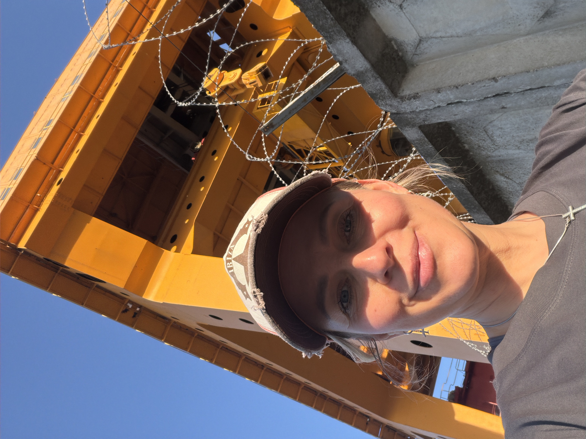 A selfie of a woman in front of a large yellow crane at an unknown location in Chaikovsky, Russia. The woman is wearing a gray T-shirt, a baseball cap, and a necklace. The crane is partially visible in the background, with barbed wire hanging from it. The sky is blue and clear. The image is slightly rotated.