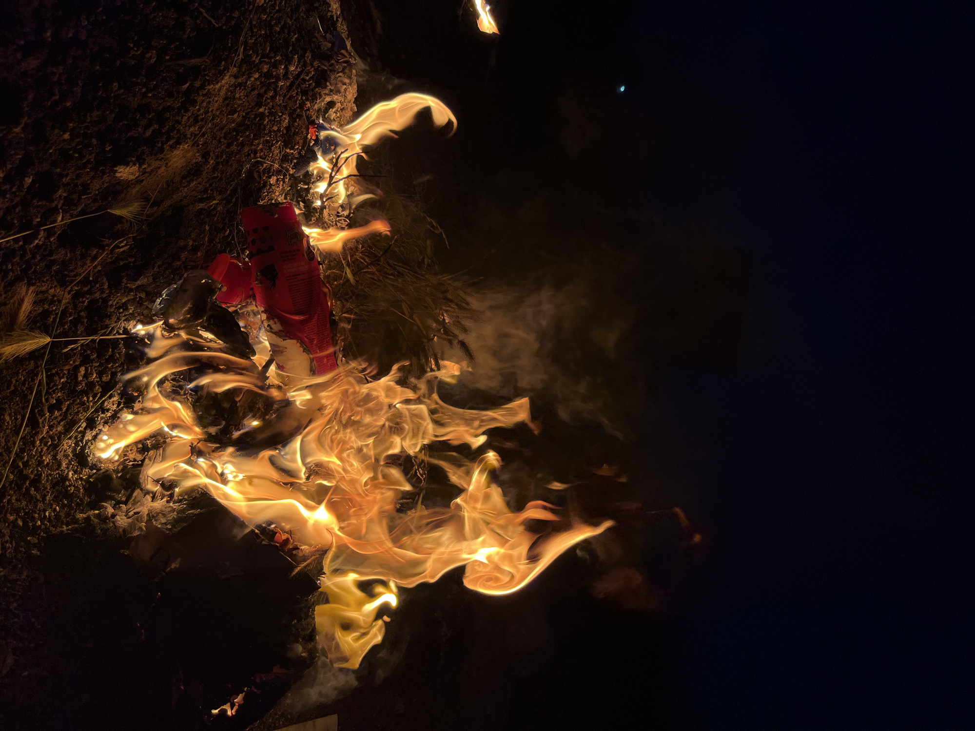 The image shows a fire burning at night, illuminating a patch of ground with dry grass and a red object, possibly a package or container, engulfed in flames. The right side of the image is dark, suggesting a night scene. The location, Kaspiysk, Russia, is provided only as background information and is not directly discernible from the image itself.