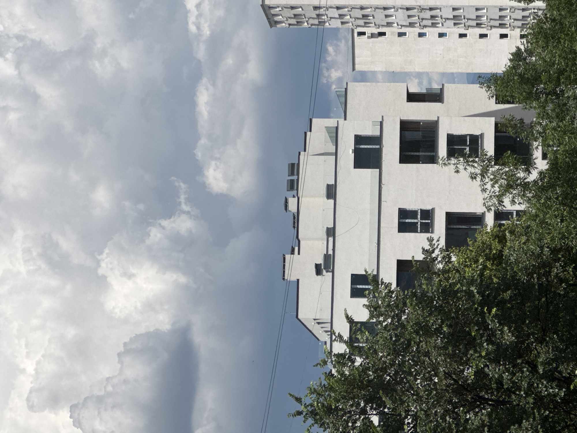 The image depicts a contemporary residential building with a white facade, characterized by numerous windows and balconies, set against a cloudy sky. In the foreground, lush green trees obscure parts of the building. To the right, a taller, similar building is partially visible, also featuring a façade with many windows. The scene appears to be in an urban setting, possibly residential. Based on metadata, this image was taken on a sunny day in Sofia, Bulgaria.
