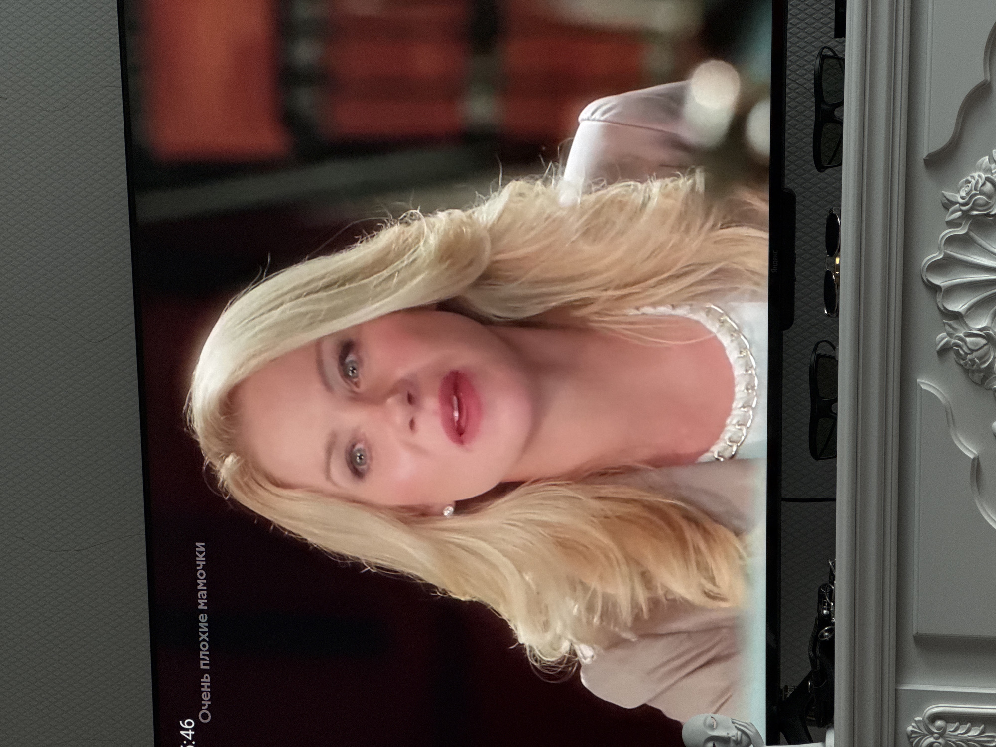 The image shows a television screen displaying a close-up of a woman with blonde hair and red lipstick. Text in Russian appears in the top left corner of the screen, reading "5:46 Очень плохие мамочки," which translates to "5:46 Very Bad Moms." The woman is wearing a light-colored blazer and a pearl necklace. The background of the screen is blurred, showing what appears to be shelves with books. Below the television, a white mantelpiece is visible, with several objects placed on it, including a small statue of a face, a toy car, and pairs of sunglasses. The overall scene is an indoor setting, likely a living room, with no discernible connection to the city of Oskemen, Kazakhstan.
