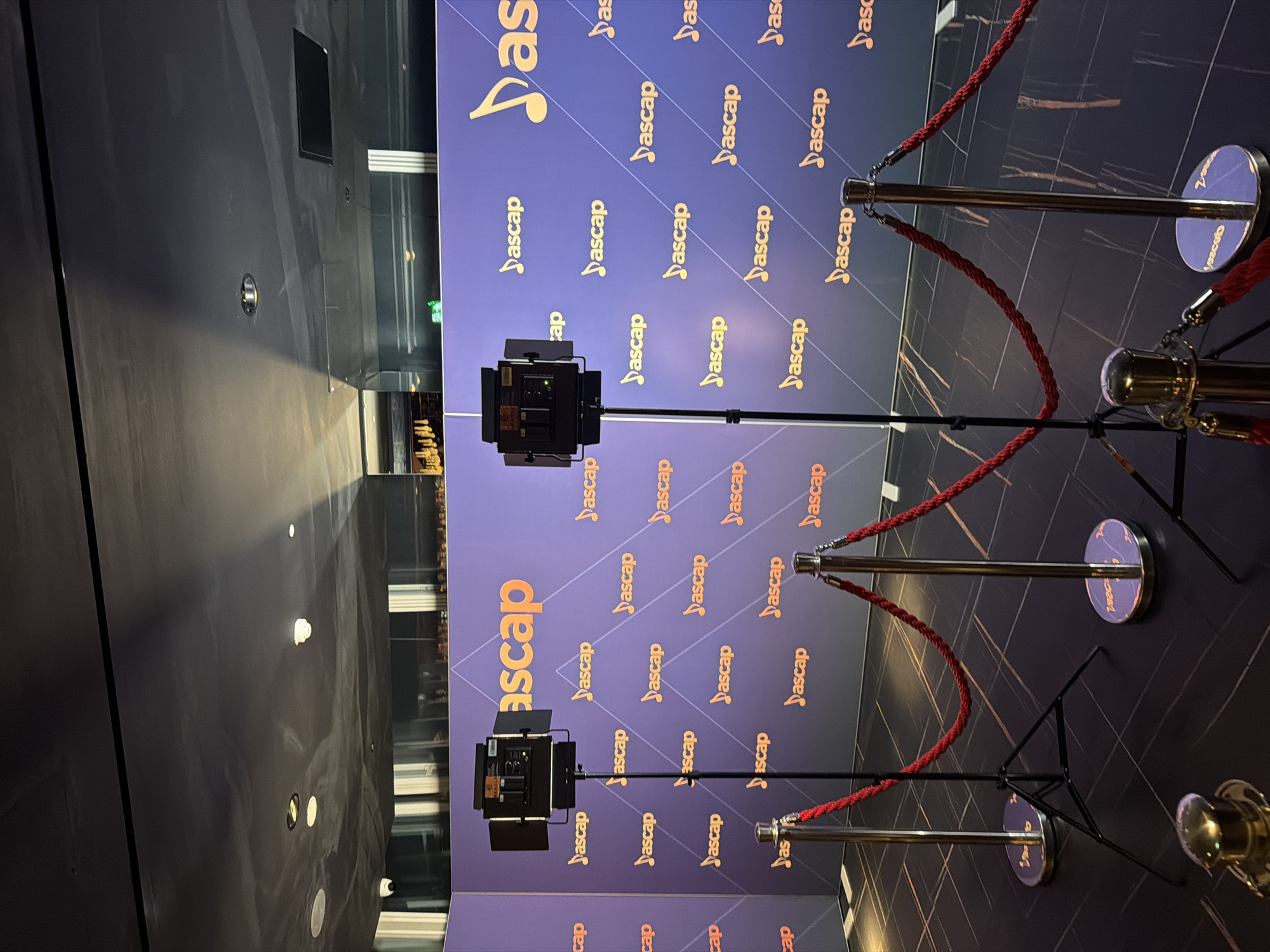 The image depicts a red carpet event setup in what appears to be an indoor venue, possibly a convention center or ballroom, in London, United Kingdom. The backdrop is a dark blue wall patterned with repeating "ASCAP" logos and musical notes in a lighter gold color. Two professional lighting rigs on tripods are positioned in front of the backdrop, suggesting a photo opportunity area. Red velvet ropes are strung between stanchions, creating a pathway. The floor is dark and reflective, with a subtle striped pattern. The lighting is dim, with spotlights from the rigs illuminating the foreground and the backdrop. No people are visible in the image.