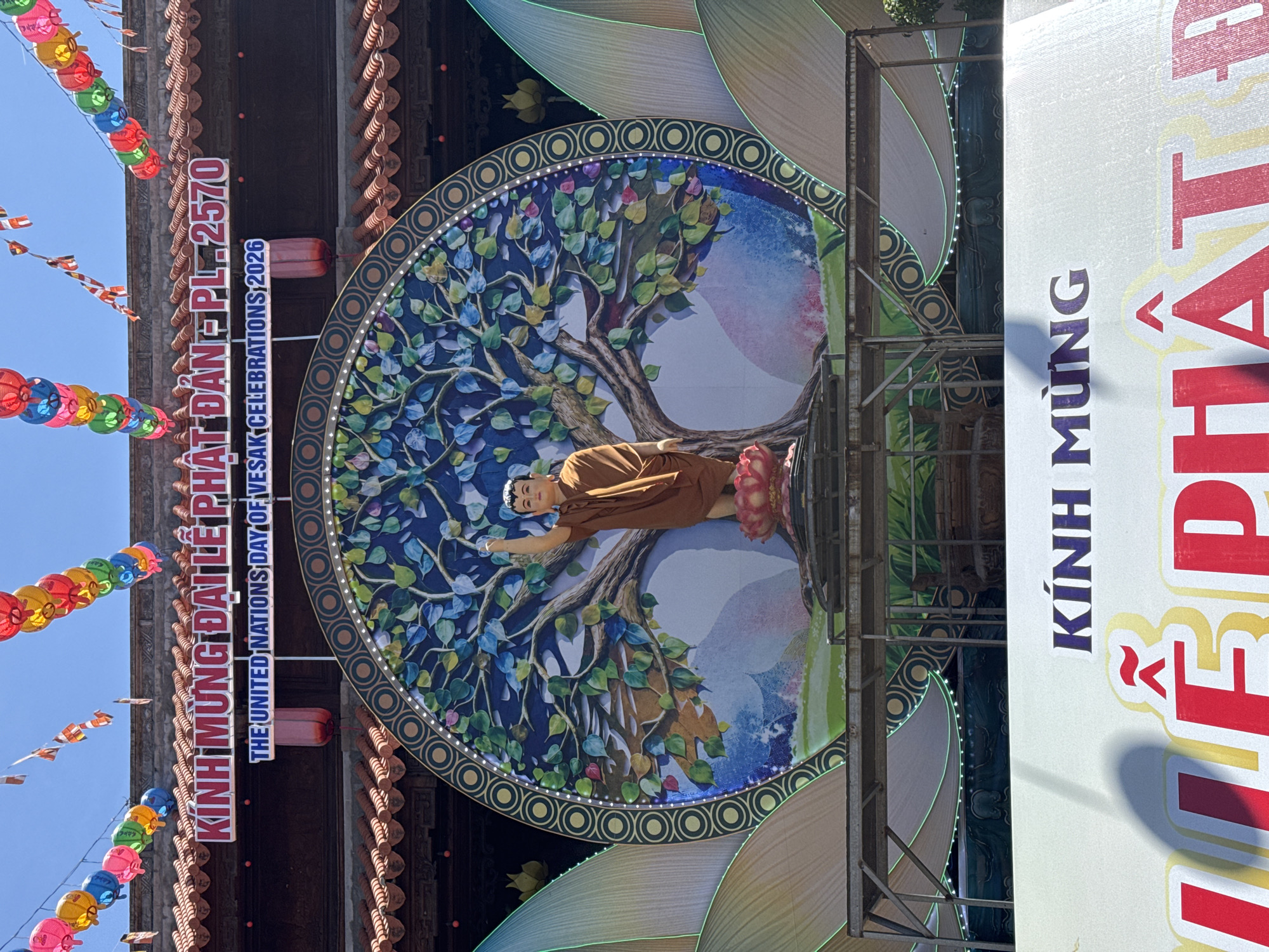Here is a detailed description of the image:
**Location:** Tay Nha Trang Ward, Vietnam.
**Scene:** An outdoor display setup, likely for a Buddhist celebration.
**Subjects and Objects:**
* A central circular backdrop depicts a tree with blue and green leaves, a figure of Buddha reclined beneath it.
* A large sign reads "KÍNH MỪNG ĐẠI LỄ PHẬT ĐẢN PL.2570." The sign includes the English text "THE UNITED NATIONS DAY OF VESAK CELEBRATIONS, 2026."
* A separate, larger sign on the right shows "KÍNH MÙNG" with the bottom words cut out of the image.
* Overhead, a string of colorful lanterns is visible.
* Scaffolding is on the right side of the display.
**Actions and Interactions:** None evident, the scene is a static display.
**Environment:** The scene appears to be set during the daytime, with bright sunlight. The presence of lanterns indicates a festive or celebratory atmosphere. The building background suggests this is an urban temple or related setting.