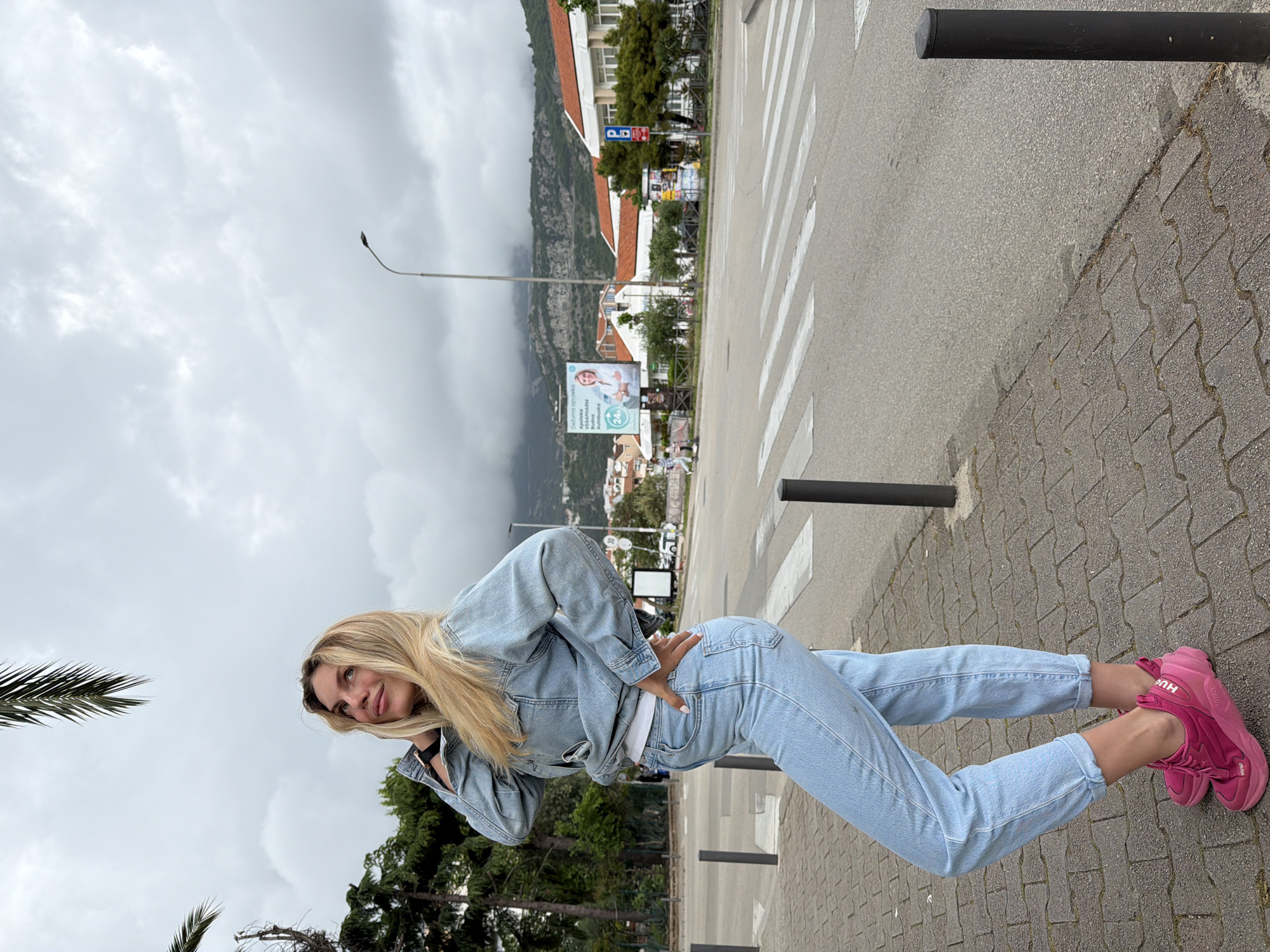 A woman is standing on a sidewalk next to a crosswalk in Budva, Montenegro. She is wearing a light blue denim jacket and jeans, a white top, and bright pink sneakers. She has long, blonde hair. Her right hand is on her hip.
The background includes buildings, trees, and a mountain partially obscured by clouds. A billboard is visible in the background. The weather appears overcast. Metal bollards line the edge of the sidewalk. The crosswalk is marked with white stripes. The sidewalk is paved with grey interlocking pavers. The road is asphalt.