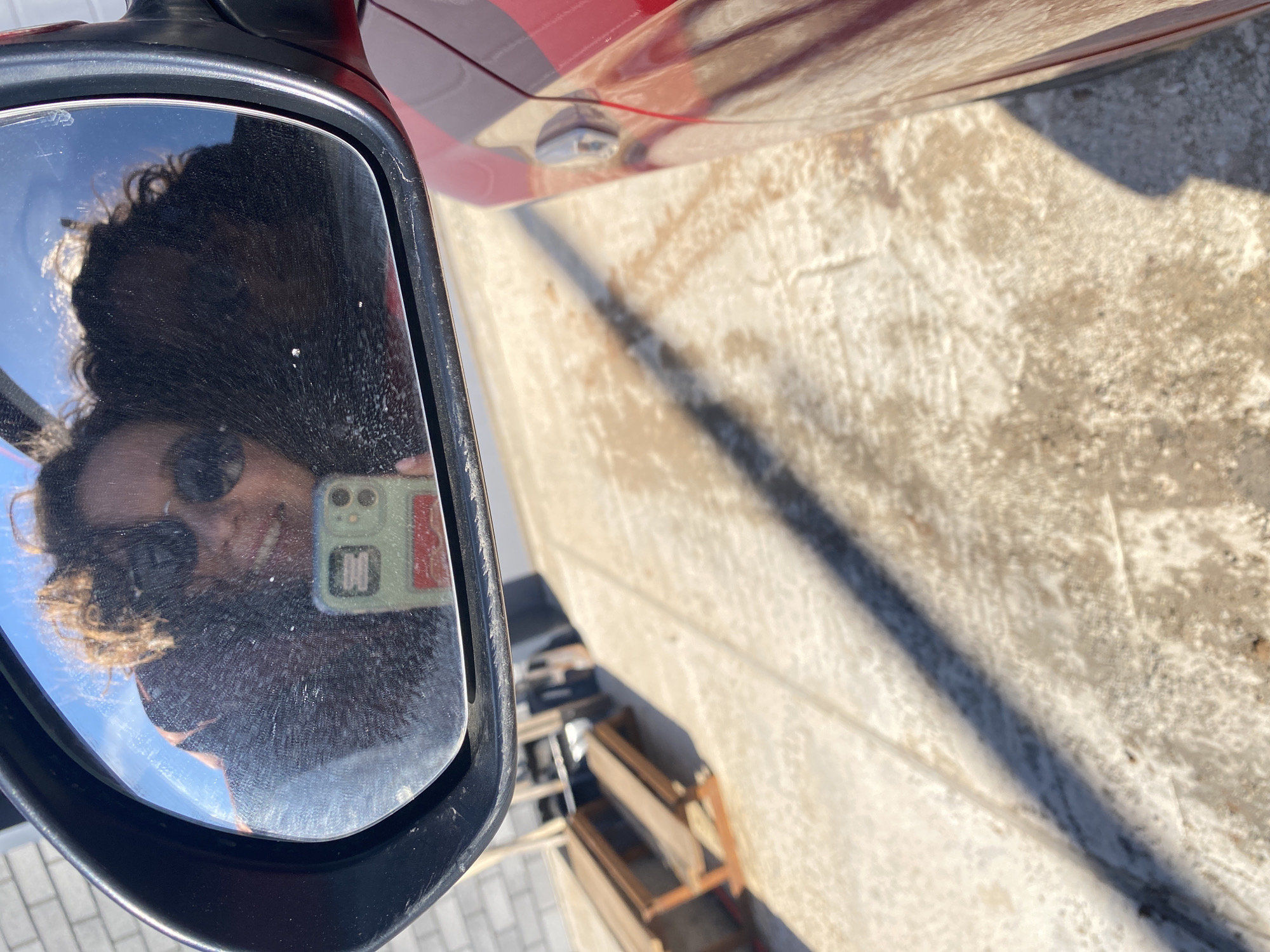 A woman with curly hair is reflected in a car's side mirror, taking a selfie with a green phone case. The car is red, and the surrounding area appears to be a sunlit, paved surface with some shadows. A wooden structure is partially visible in the background. The location is most likely in a city, possibly Ольховка, Russia.