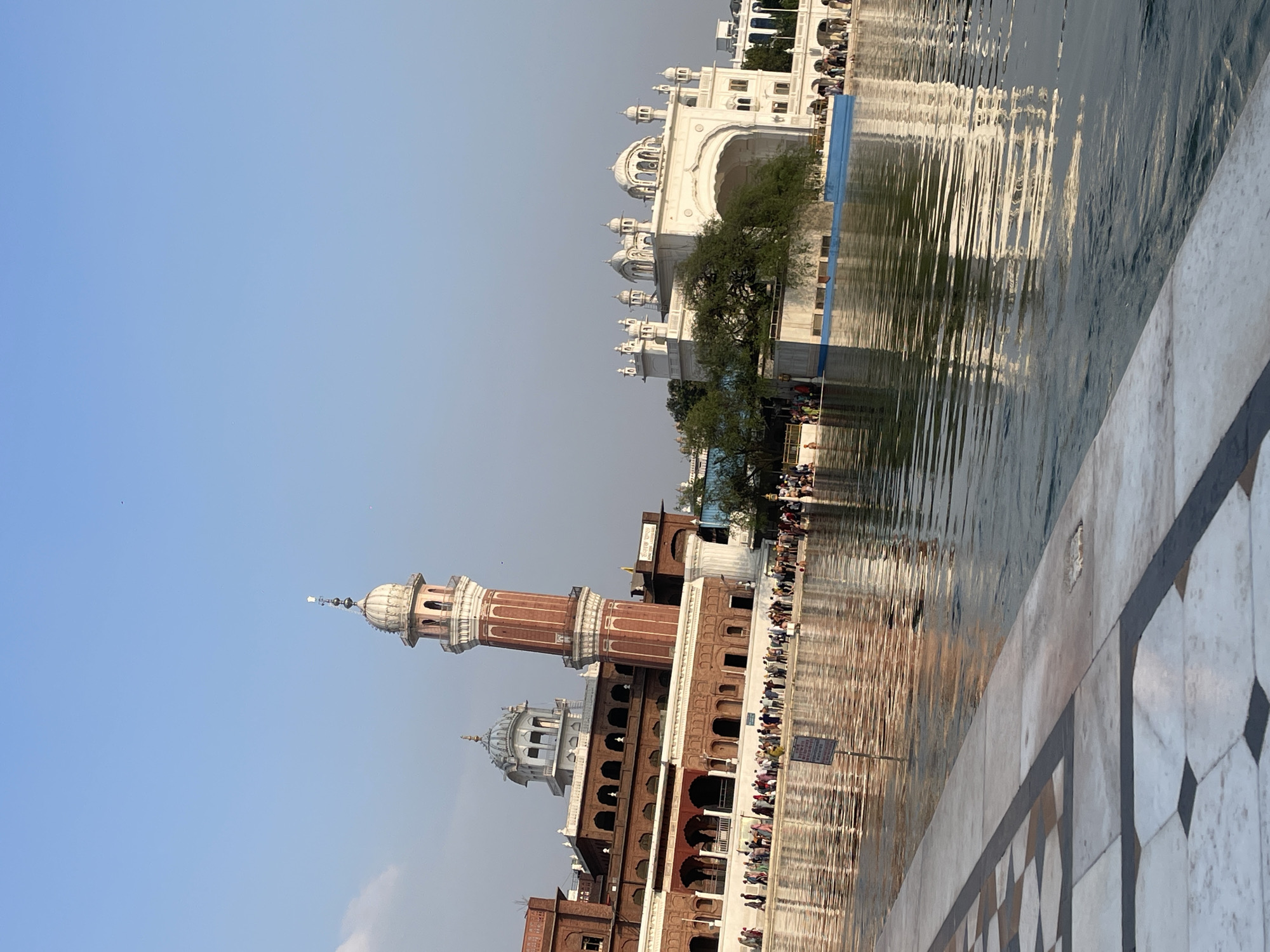 The image depicts the Golden Temple complex in Amritsar, India. The scene is dominated by the reflective waters of the sarovar (pool) in the foreground, with the iconic architecture of the temple and its surrounding buildings visible across the water.
In the background, a tall, red and white striped minaret pierces the clear blue sky, a prominent feature of the complex. Adjacent to it, a building with arched windows and a white dome is visible. To the right, a grand white structure with multiple domes and arches stands out, reflecting in the water. A lush green tree partially obscures this white building.
A large number of people are gathered along the edge of the sarovar, some standing, others appearing to be bathing or walking. Their presence suggests a pilgrimage or a visit to this sacred site. The water itself is calm, with ripples creating a distorted reflection of the surrounding architecture. The foreground features a patterned marble walkway, likely part of the perimeter of the sarovar.
The overall impression is one of serenity and spiritual significance, typical of the Golden Temple. The clear sky and the calm water contribute to a peaceful atmosphere.