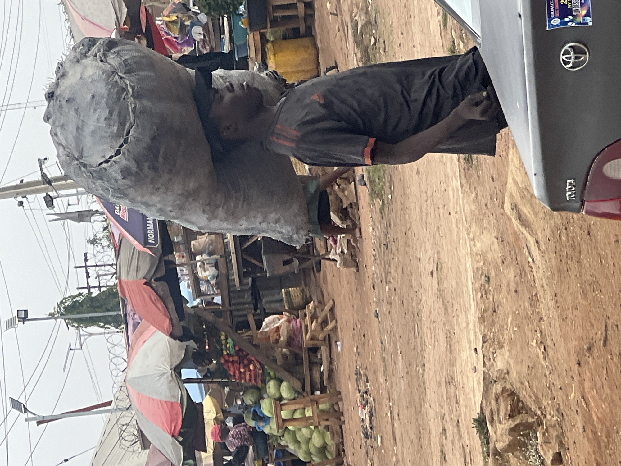 A man carries a large, heavy-looking sack on his left shoulder in Gura Top, Nigeria. He is wearing a dark-colored t-shirt and dark shorts and leans slightly to his right to counterbalance the weight of the load. He is standing near the open trunk of a gray Toyota car. The scene unfolds in what appears to be a bustling market area with several stalls selling fruits and vegetables. Colorful umbrellas and canopies provide shade, and overhead electrical lines are visible. The ground is dirt and appears somewhat uneven. People are walking around in the background.