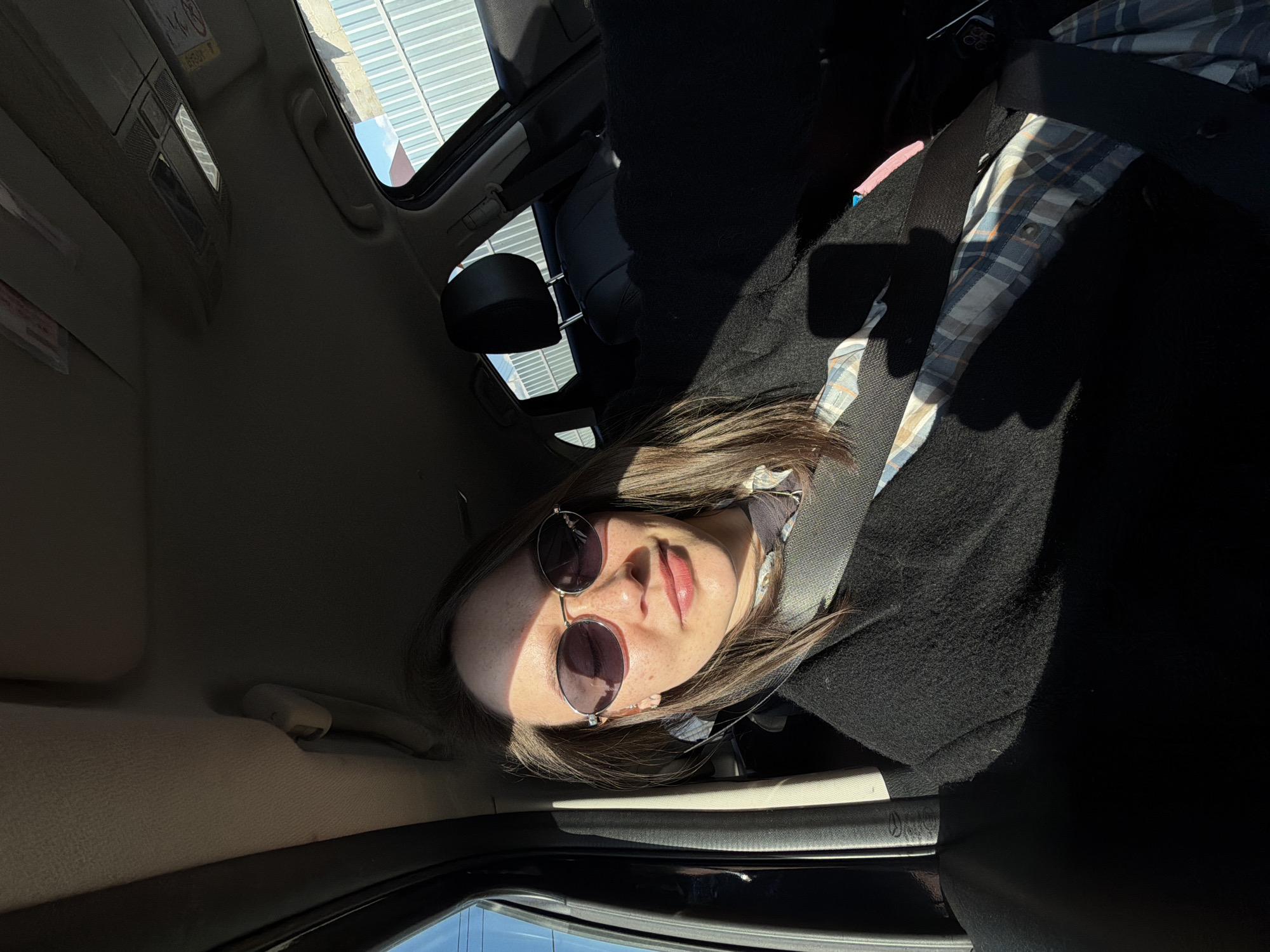 A woman wearing sunglasses is inside a car. The location appears to be Chaikovsky, Russia. The woman is wearing a black coat and a plaid shirt. She is wearing a seatbelt. The car is parked near a building with a metal roof. The image is taken from an unusual angle.