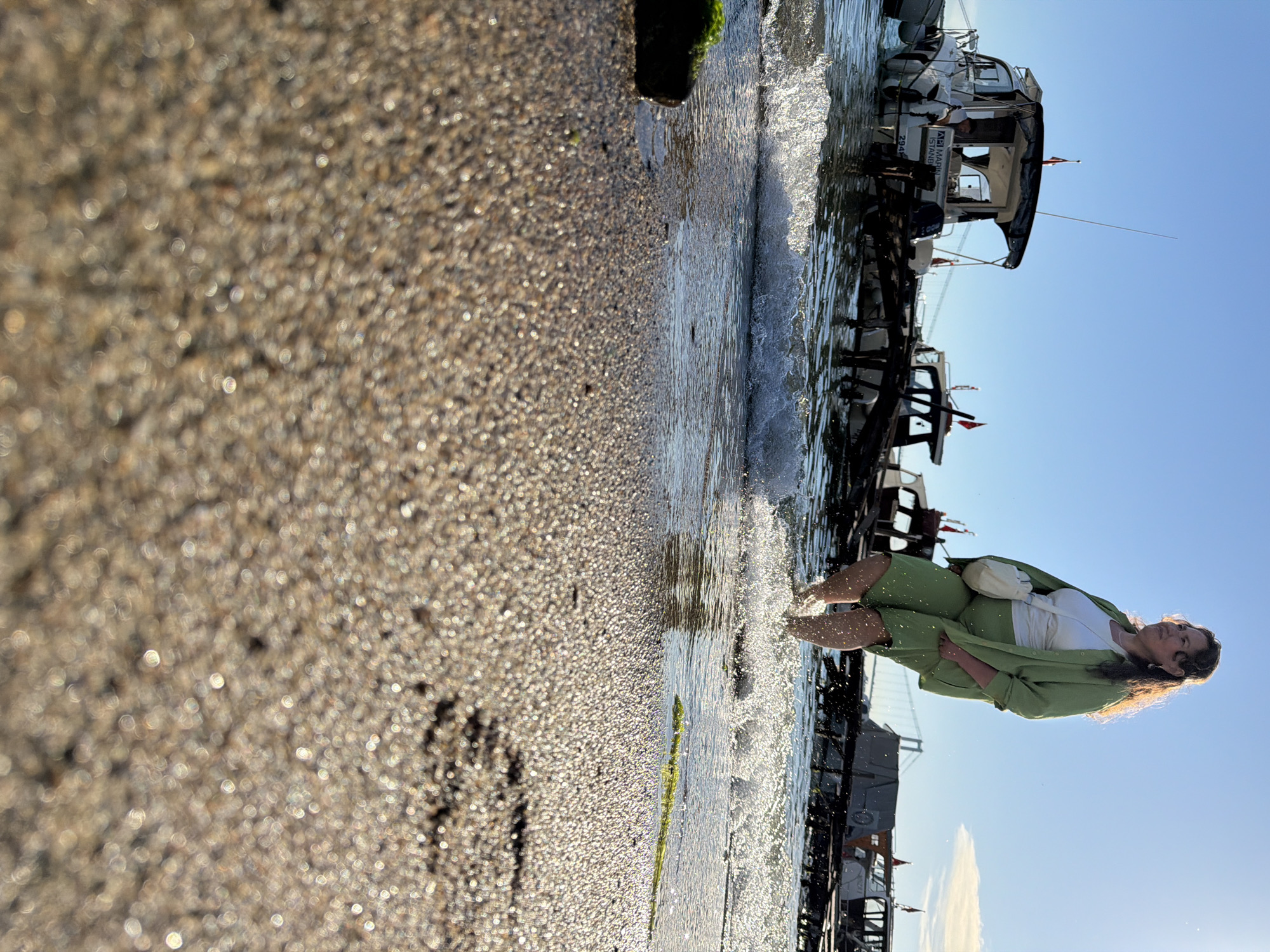 The image captures a woman walking through shallow water near a beach in Üsküdar, Turkey. She is wearing a light green suit and a white top, with a cream-colored bag slung over her shoulder. Her feet are splashing in the water, creating ripples. In the background, several boats are docked near a wooden pier. The sky is clear and blue, with the sun setting, casting a warm glow on the scene. The foreground is dominated by a close-up view of wet sand, reflecting the sunlight.