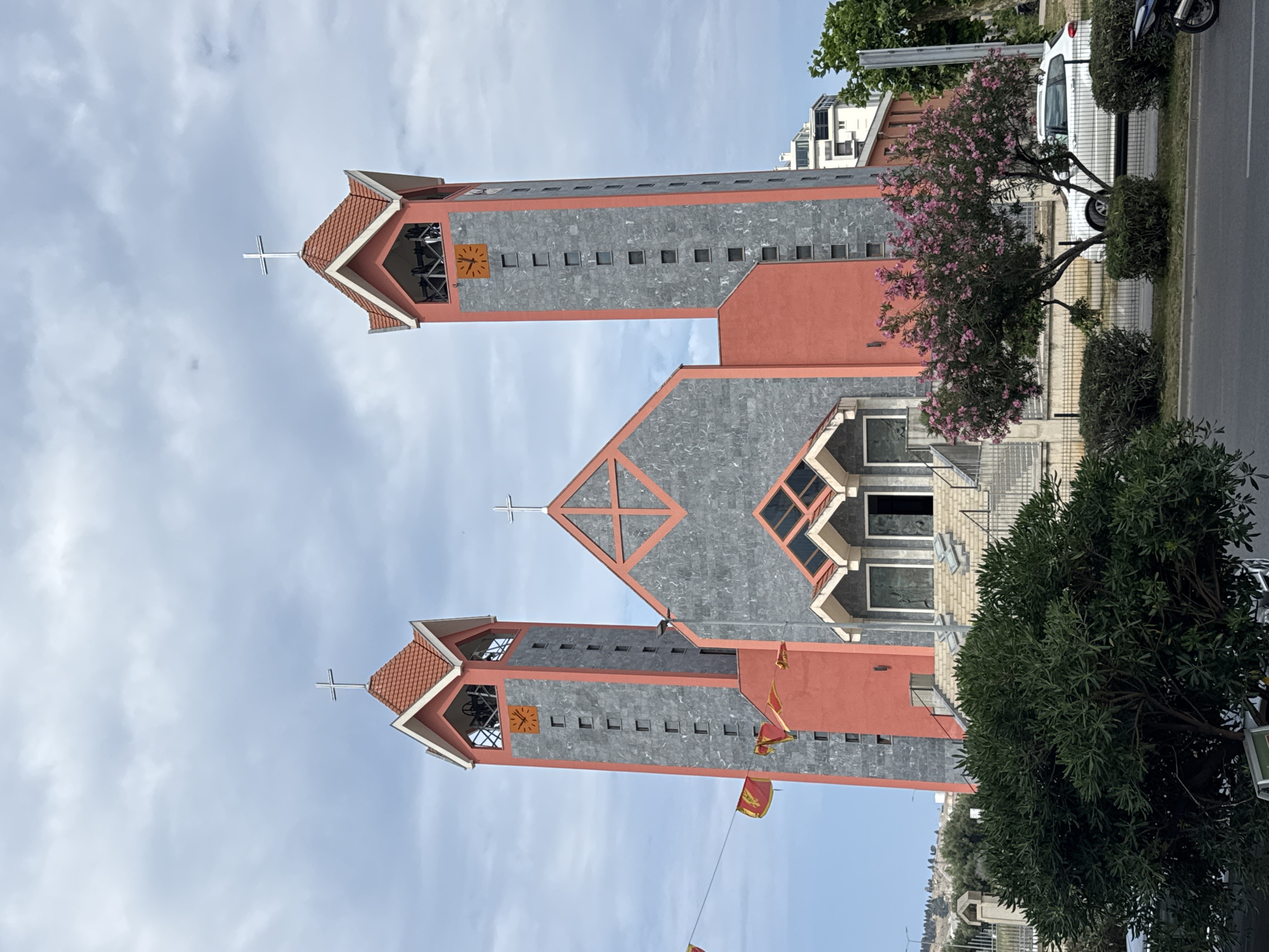 This is a low-angle, medium shot of the Church of the Holy Prophet John the Baptist in Bar, Montenegro, captured on a cloudy day. The church features a prominent central section and two tall, slender bell towers on either side. The central part of the facade is clad in gray stone with orange geometric accents, topped by a red-tiled, angular roof. The bell towers are primarily red, with gray stone sections on their sides. Each tower has a clock face with orange numerals. The main entrance is approached by a wide set of stairs. In front of the church, there are green bushes, a tree with pink blossoms, and a black metal fence. A white car is parked on the street in front of the fence. Several red and yellow flags are strung across wires in the foreground. The sky is overcast with scattered clouds.