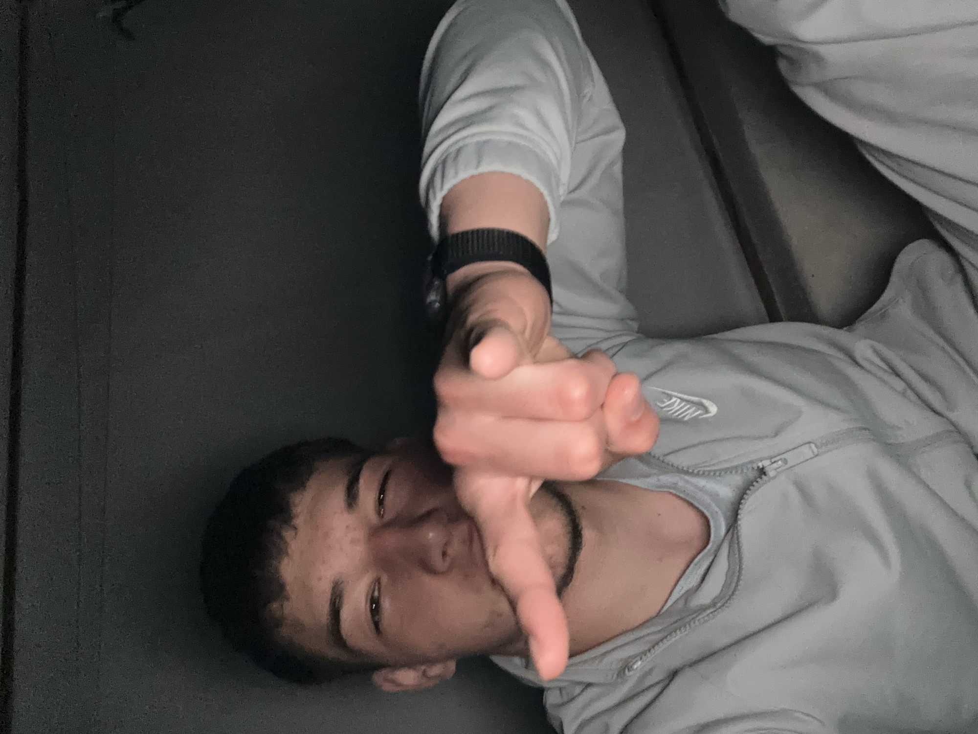 A person with short dark hair is lying down and giving a thumbs down gesture towards the camera. The person is wearing a light grey Nike zip-up jacket and a black wristband. The background appears to be a dark grey or black surface, possibly a wall or fabric. The image is poorly lit. The city and country are not discernible from the provided image.