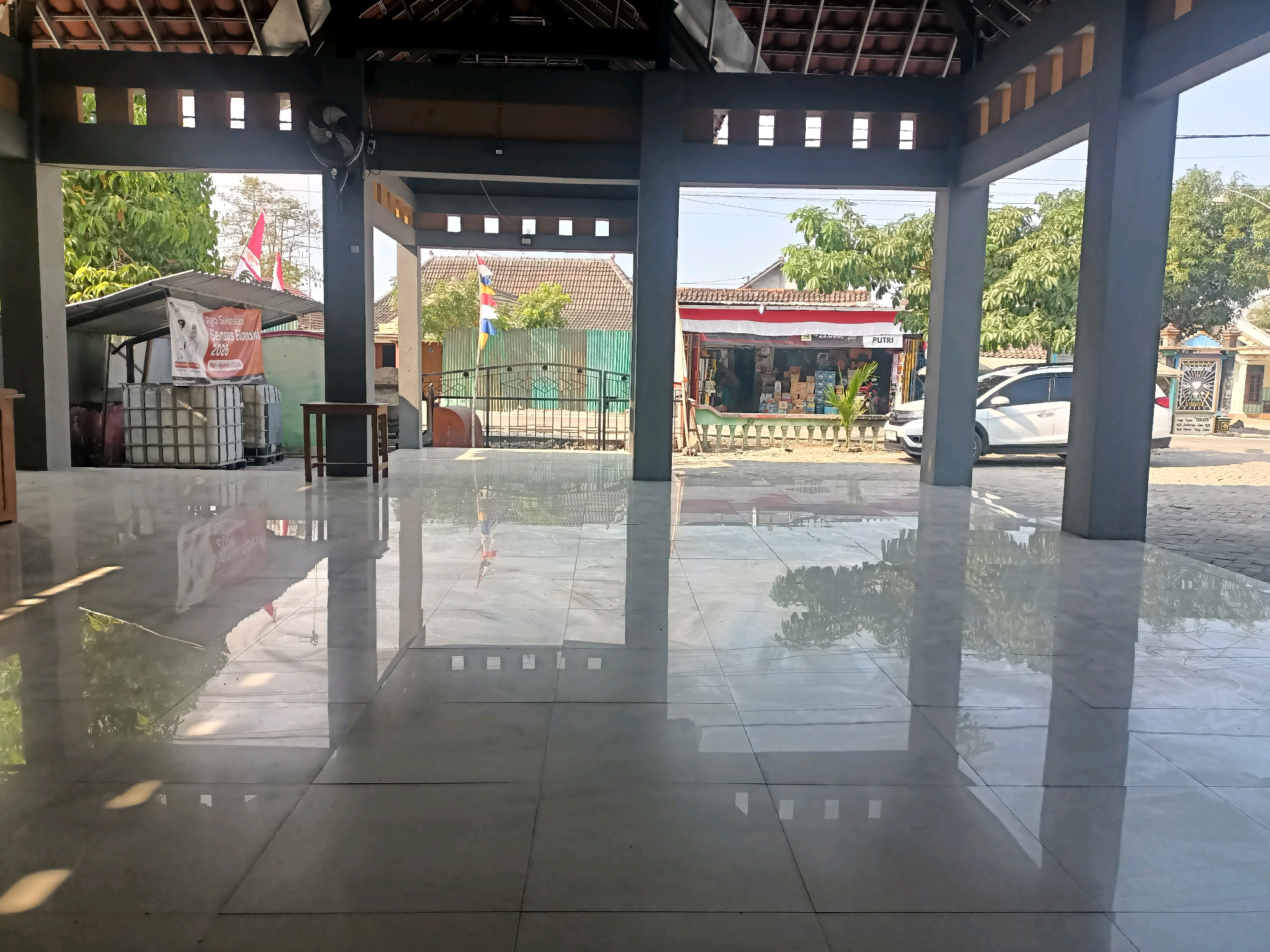 The image captures a view from inside a covered, open-air structure with a tiled floor and a partially visible roof. The floor is highly reflective, mirroring the surroundings. Through the open structure, a street scene in Sumber Bendo, Indonesia, is visible. On the left, there is a small market stall with stacks of white containers and a banner. Further down the street, a red and white awning stretches across a building, likely a shop. A white car is parked on the right side of the street. Trees and houses with tiled roofs are scattered throughout the background. Flags, including Indonesian flags, are also visible. The overall impression is a bright, sunny day in a rural or semi-urban Indonesian setting.