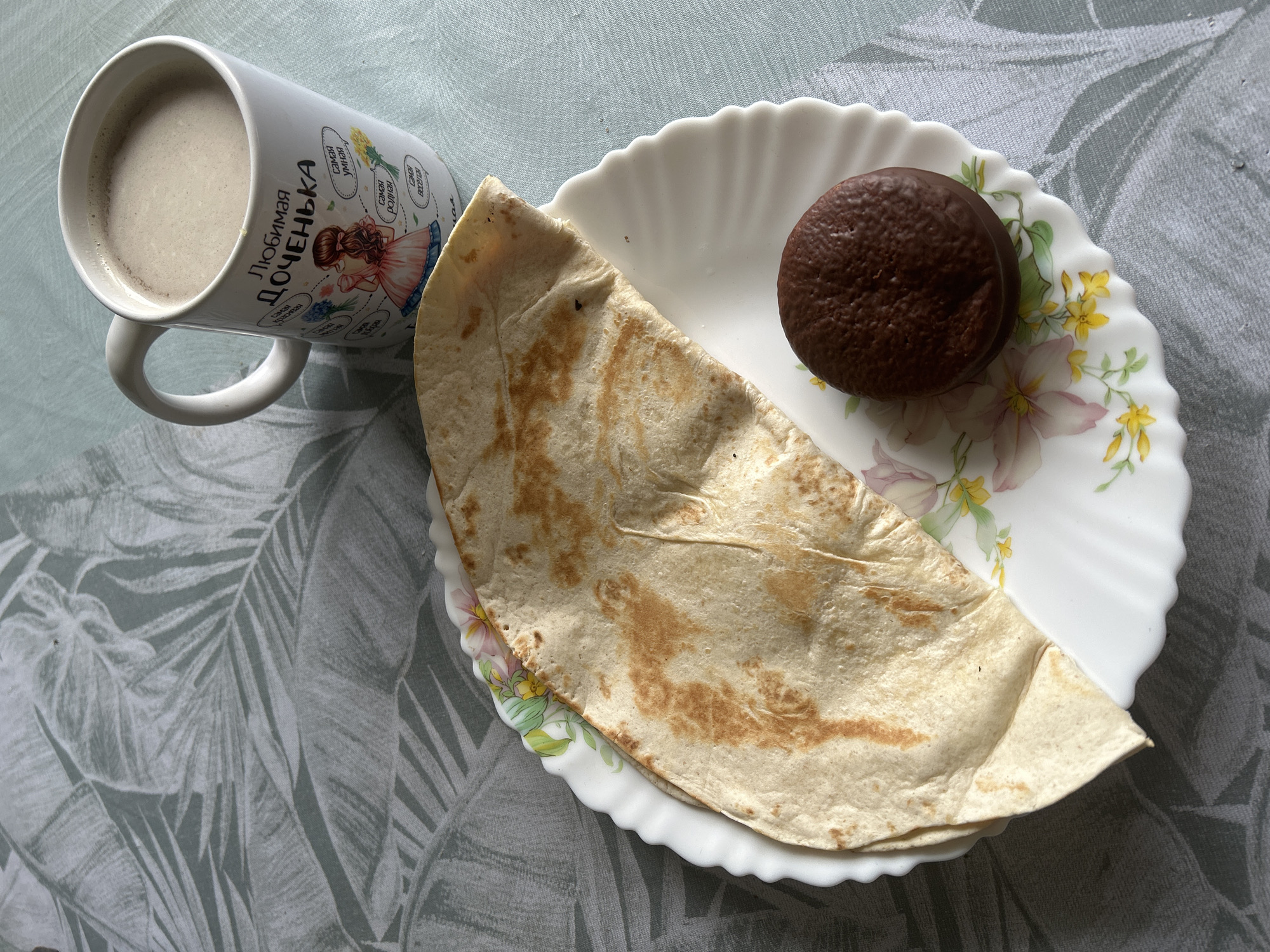 A top-down shot shows a mug, a plate, a flatbread, and a chocolate snack item arranged on a surface with a leaf-patterned cloth.
The mug is white with Cyrillic text and illustrations, filled with a light brown beverage.
The plate is white with a scalloped edge and floral decorations. The flatbread is folded and occupies a large portion of the plate. A round chocolate snack item sits to the side of the flatbread.
The background is a light green cloth featuring a pattern of broad, stylized leaves.
The location in the city of Фоки, Russia is not discernible from the provided image.