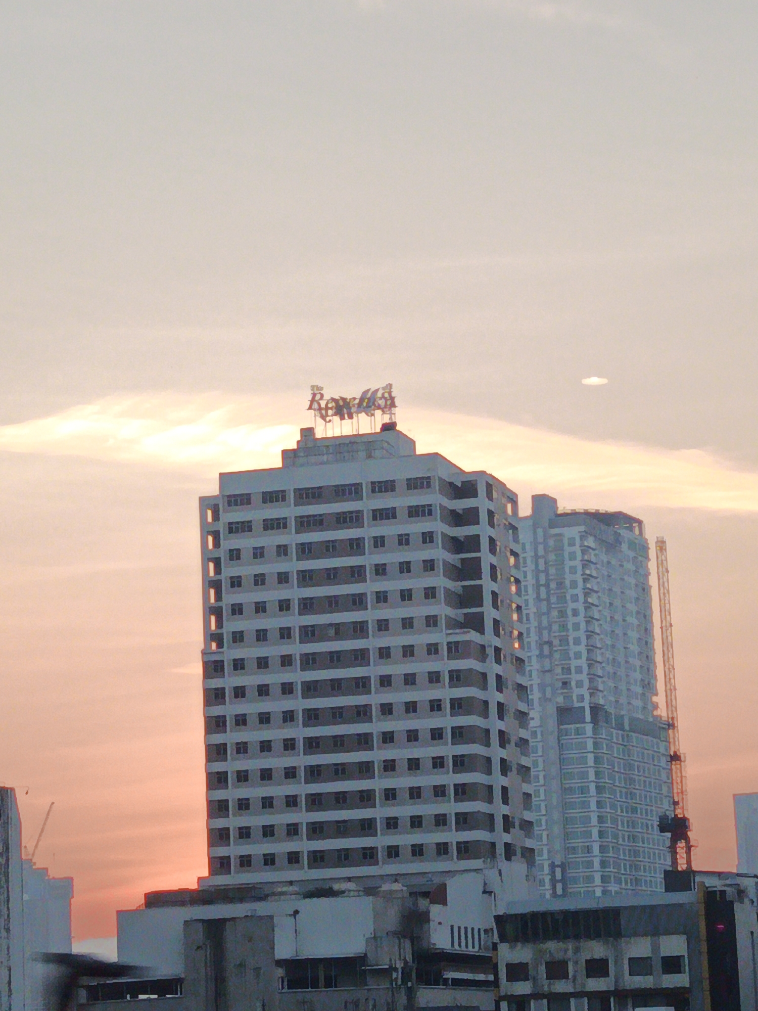 An outdoor, eye-level, medium shot captures a cityscape at dusk. The sky is a pale orange, transitioning to a lighter hue towards the top. In the foreground, two large, multi-story buildings dominate the frame. The building on the left is lighter in color and has a sign with illuminated lettering at the top that reads "The Rexkl." The building on the right is taller and a darker shade of blue-gray, with a lattice-like pattern on its facade. A tall construction crane with a red base stands to the right of the darker building. Other, lower buildings and structures are visible in the bottom portion of the image, some appearing under construction. The overall impression is one of urban development against a soft, twilight sky.