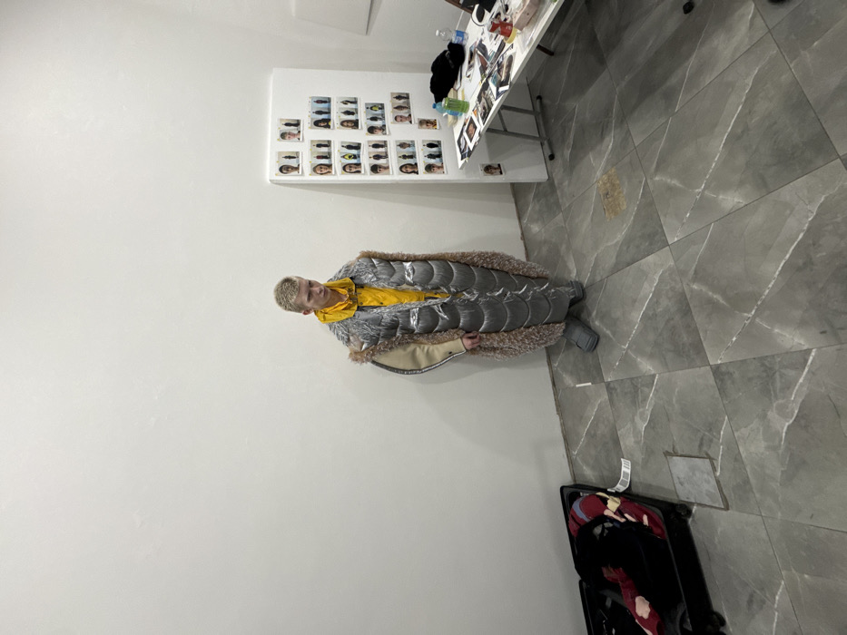 The image depicts an indoor scene, likely a backstage or studio area for a fashion event in Milan, Italy. A young individual, centrally positioned and facing slightly right, has short, light-colored hair and wears a bright yellow long-sleeved top. Over this, they wear a prominent, floor-length garment featuring a silver, quilted puffer-style main body with a substantial, light brown/beige furry trim down the front and around the hem. A lighter beige fabric panel is visible on the upper left arm. The individual wears large, light gray, slide-style footwear. To the left of the individual, a black open suitcase contains folded clothing, including a red item. To the right, a white board propped against the wall displays multiple small portrait and full-body photographs of various individuals, arranged in columns. Adjacent to the board, a white folding table is laden with papers, magazines, two bottles (one green, one clear), and dark fabric items. The room features plain white walls and a grey tiled floor with a subtle marble pattern. The lighting is even and appears artificial.