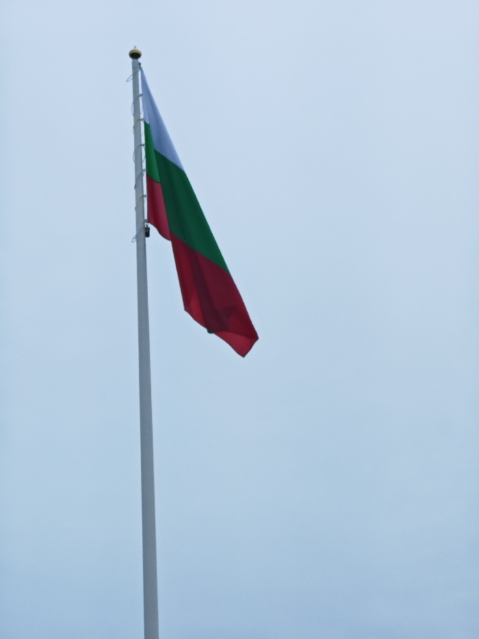 A Bulgarian national flag is visible, partially unfurled, against an overcast light blue-gray sky. The flag displays horizontal stripes of white, green, and red from top to bottom. It is attached to a tall, white pole that extends vertically from the bottom edge of the frame towards the upper left. A golden spherical finial tops the flagpole, and hoist ropes are visible running down the pole's side. The flag is positioned in the upper-left quadrant of the frame, angled downwards to the right, indicating a gentle breeze. The scene is located in the city of Shumen, Bulgaria, as identified by the national flag. No people or other objects are visible.