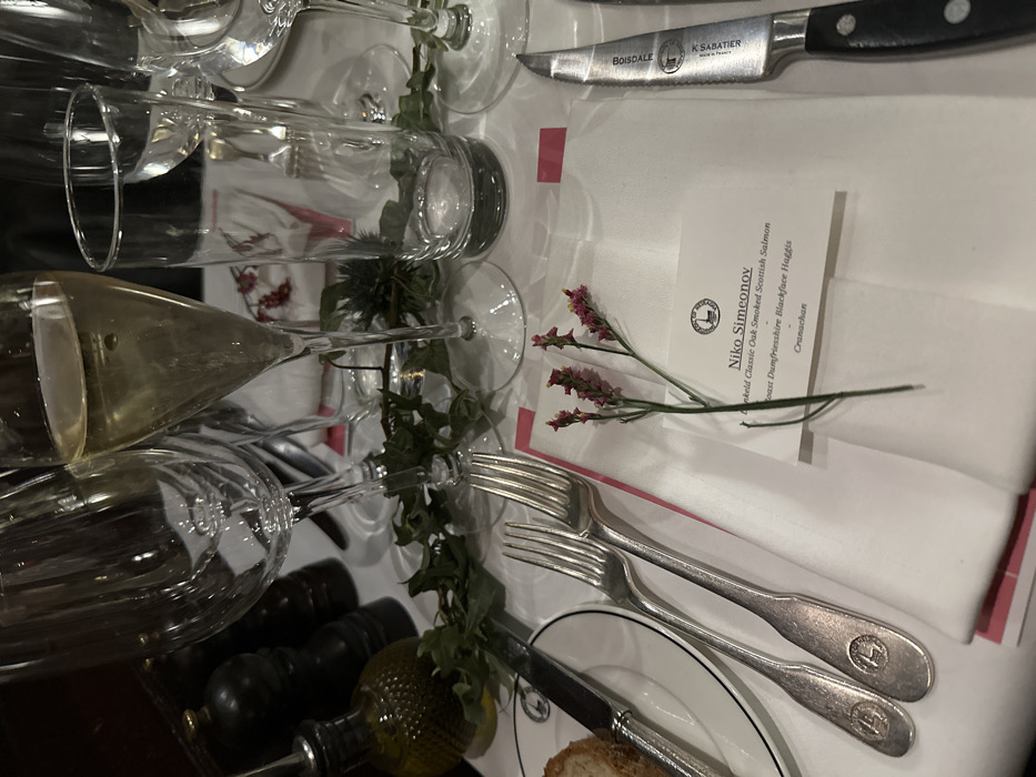 A formal table setting displays crystal glassware and silver cutlery. A menu card reads "Niko Simeonov" with the menu items "Dunkeld Classic Oak Smoked Scottish Salmon," "Roast Dumfriesshire Blackface Haggis," and "Cranachan." A single sprig with delicate pink flowers rests on the menu card. A knife with a black handle and serrated blade, labeled "Boisdale" and "K Sabatier," lies above the menu. The table is adorned with a sprig of greenery, a black pepper grinder, and olive oil. A white plate with a black rim contains a piece of bread.