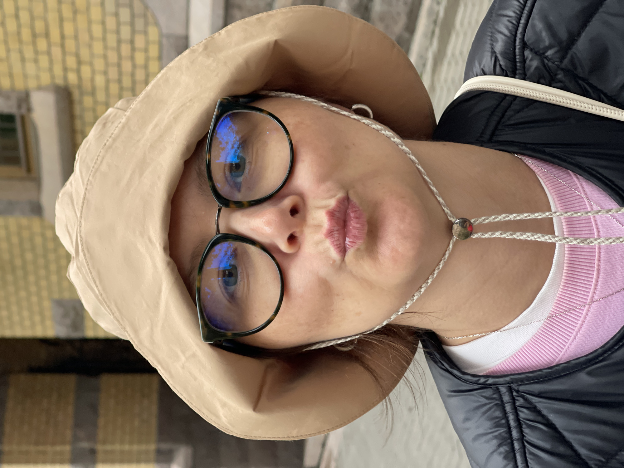 A close-up, frontal shot shows a woman wearing a beige bucket hat and a black puffer vest over a pink sweater and a white undershirt. She has on large, tortoiseshell-framed glasses. Her lips are puckered as if for a kiss. A braided cord from the hat is visible around her neck, fastened with a silver button. A thin silver necklace is also visible. A small silver hoop earring is on her left ear. The background appears to be an exterior setting with a yellow brick building and some muted colors suggesting a city environment, potentially Saint Petersburg, Russia.
