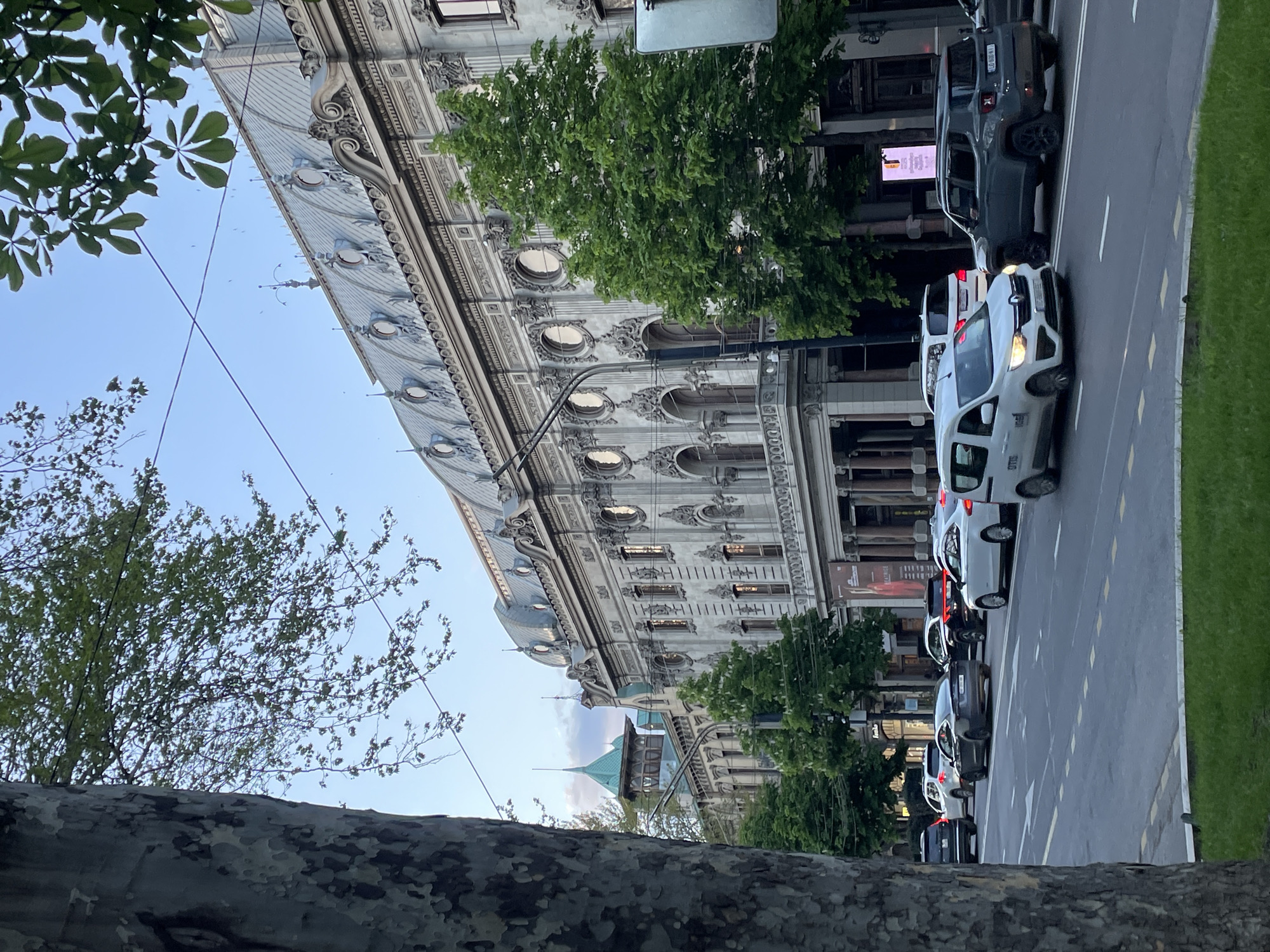 This media file captures a vibrant street scene in Tbilisi, Georgia, during what appears to be the late afternoon or early evening, under a clear, bright blue sky.
The central focus is a magnificent, multi-story building with a light-colored, highly ornate facade, adorned with classical architectural elements, such as intricate moldings, pilasters, arched windows, and a distinctive series of circular attic windows (oculi) along its roofline. Behind and to the right of this grand structure, a smaller building with a prominent green-domed or turreted roof adds to the architectural landscape.
The paved street in front of the building is lined with several cars, indicating typical urban activity. Visible vehicles include multiple white cars, likely taxis or vans (one appears to be a Renault with the number "97799" on its side), and a dark grey Jeep Renegade SUV with the license plate "LO-080-AV". A grassy median or sidewalk runs alongside the street.
Green trees with lush foliage are present throughout the scene, with a large tree trunk in the bottom left foreground and leaves framing the top left, suggesting a tree-lined street. Utility wires are visible crisscrossing the sky. On the building facade, a digital billboard displays an advertisement, and a banner sign below it clearly reads "GALLERY" and "A MUSEUM", identifying the building's purpose. No people are distinctly visible in the frame, but the parked and moving cars suggest a bustling urban environment.