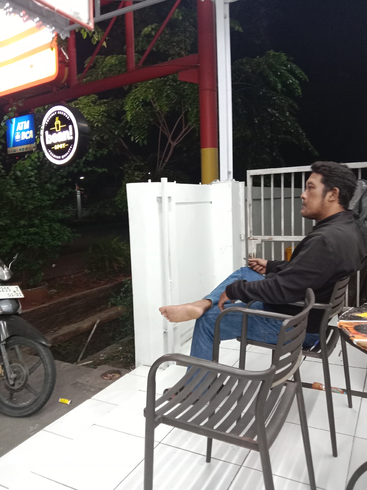 A man is sitting on a chair outside an establishment at night. He is wearing a black jacket and blue jeans. The man appears to be smoking a cigarette and is looking to his right. In front of him is a table with another chair. To his left, there is a motorcycle parked. Behind him, there is a white gate and a building. A sign for "bean! SPOT" is visible to the left of the man, which is located in Semarang, Indonesia. The ground is tiled and there are plants and trees in the background.