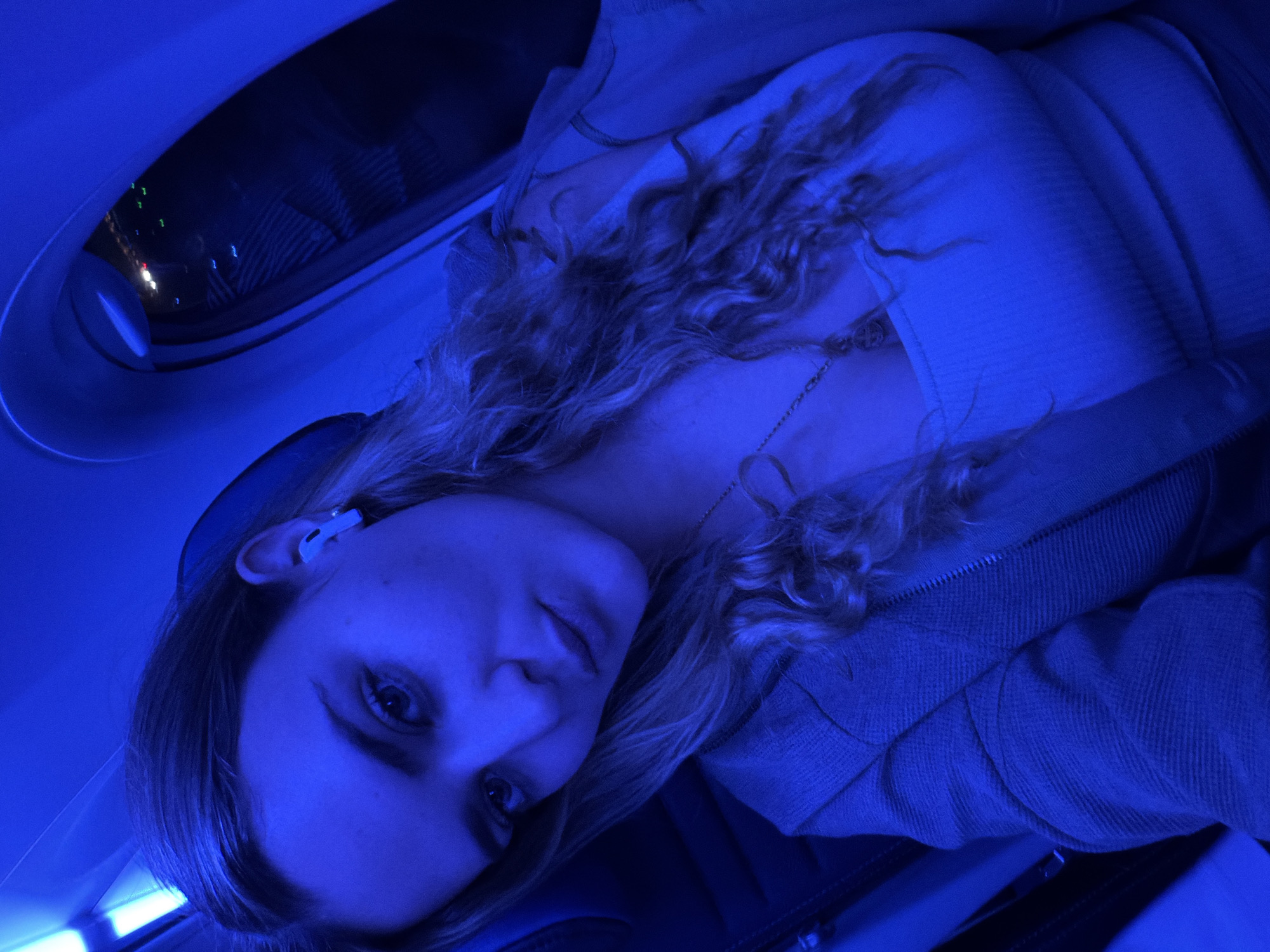 A female passenger on an airplane looks at the camera. The airplane window is on the upper left corner. The time is night because there is city lights from the window. The female is wearing a shirt and a jacket and has curly hair. She is also wearing an airpod. The location of the image could not be ascertained based solely on the image details.