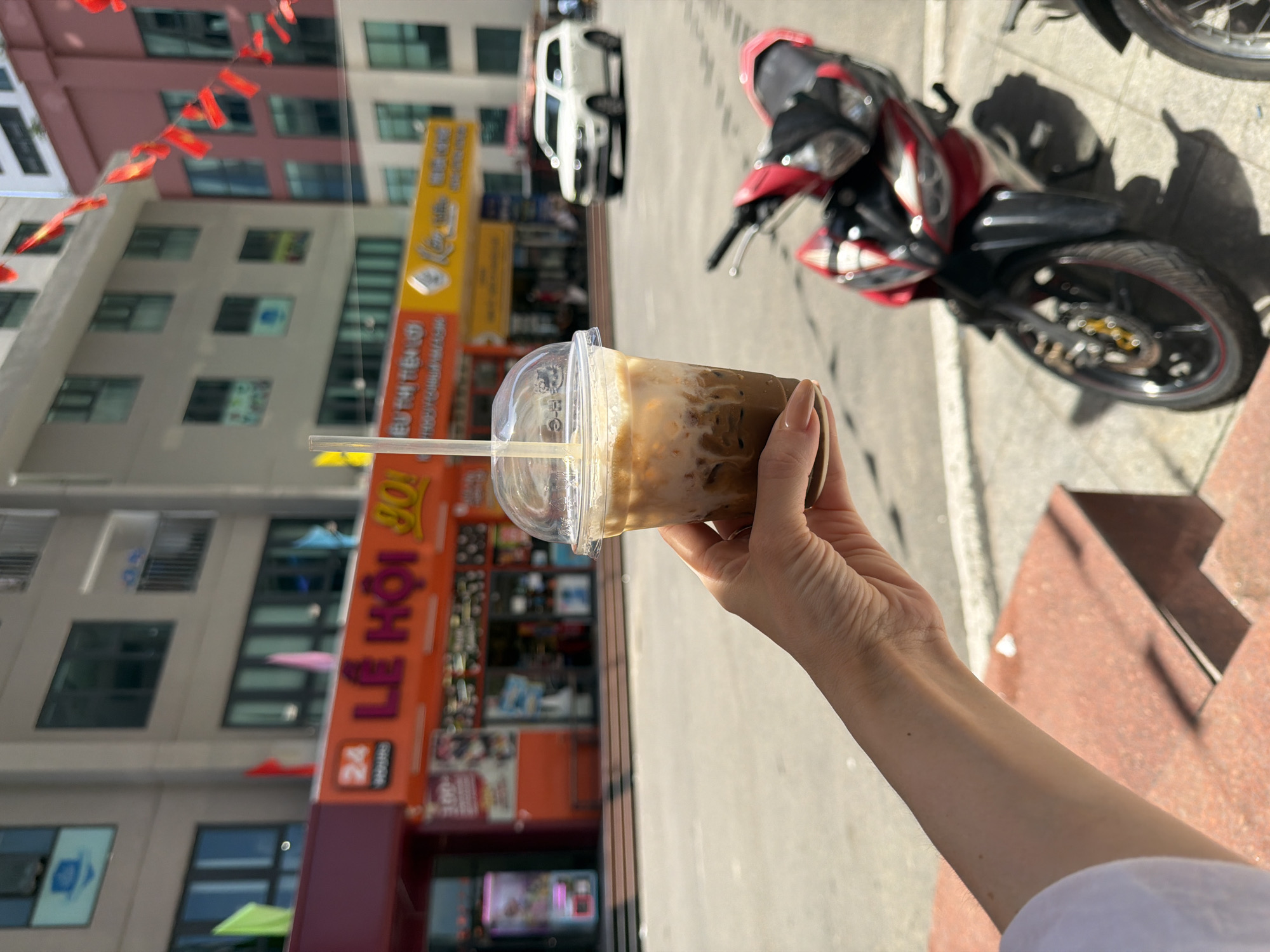 The image presents an outdoor urban scene in Bac Nha Trang Ward, Vietnam, under strong daylight conditions. In the lower-left foreground, a human right hand with light-colored manicured nails holds a clear plastic cup containing an iced beverage, layered with a dark liquid and a white creamy component, with a clear straw inserted. To the immediate right, a red and black motorcycle is partially visible, parked on a tiled sidewalk. A paved street extends into the midground, where a white sport utility vehicle is observed further down the road. The background consists of multi-story buildings. A prominent orange commercial sign on one building displays Vietnamese text, including "LỄ HỘI" (Festival) and "GO!", along with a "24 HOURS" logo. Red and various colored decorative flags are suspended above the street and along the building facades. Windows and balconies are visible on the upper levels of the background structures.