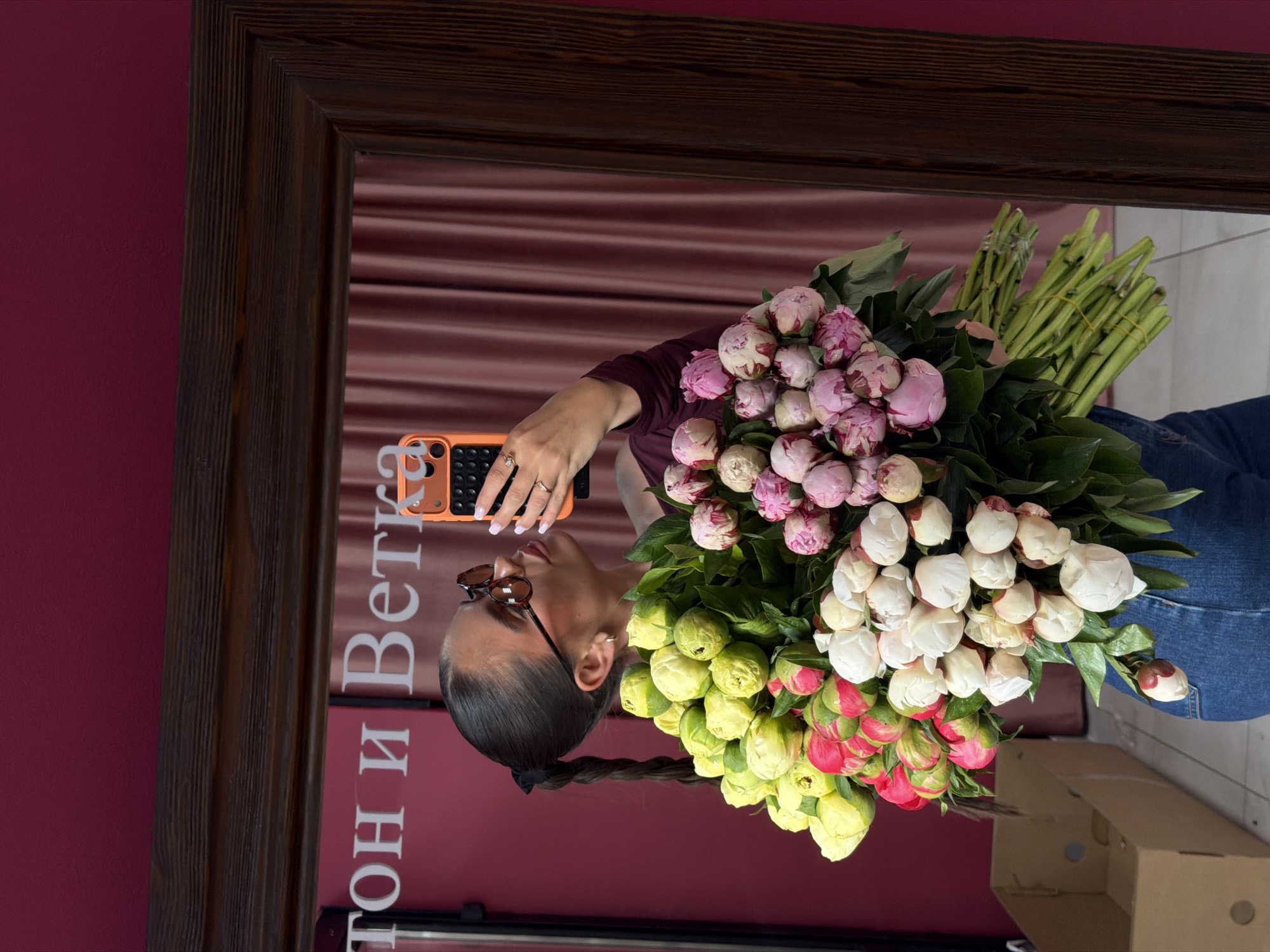 A reflection in a wooden-framed mirror shows a woman holding a bouquet of mixed-color peonies and a smartphone. The wall behind the mirror is maroon. The reflection reveals that the woman is wearing glasses, a maroon top, and jeans. Her hair is tied in a ponytail. The bouquet comprises peonies of varying colors, including pink, white, yellow, and red. The text "тон и ветка" is visible in the reflection. The city of Krasnoyarsk, Russia is implied by the text and image source.