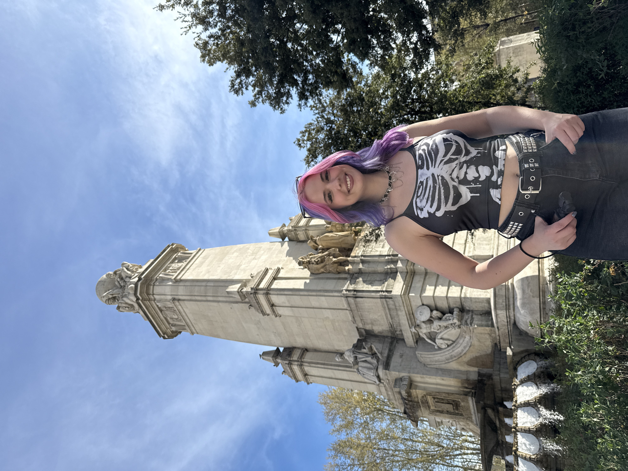 A young woman with pink and purple hair stands in front of the Monument to Cervantes in Madrid, Spain, smiling at the camera. She is wearing a skeleton-print tank top, dark jeans, and a studded belt. The monument, a large stone structure with statues, is partially visible behind her. The sky is blue with some clouds, and trees are visible in the background.
