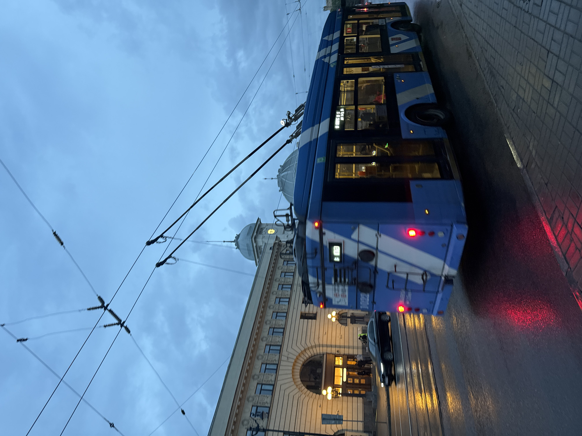 An image depicts a blue and white trolleybus driving past a beige building in Saint Petersburg, Russia. The trolleybus is in motion, indicated by the slight blur of its wheels and the streaks of light reflecting off the wet street. The building, which appears to be a train station, features an arched entrance and numerous windows. The sky is overcast, contributing to the dim lighting. Overhead, a network of trolleybus wires crisscrosses the frame. A black car is also visible on the street, driving in the same direction as the trolleybus. The sidewalk is paved with cobblestones. The overall scene suggests an early evening or a day with poor weather conditions.