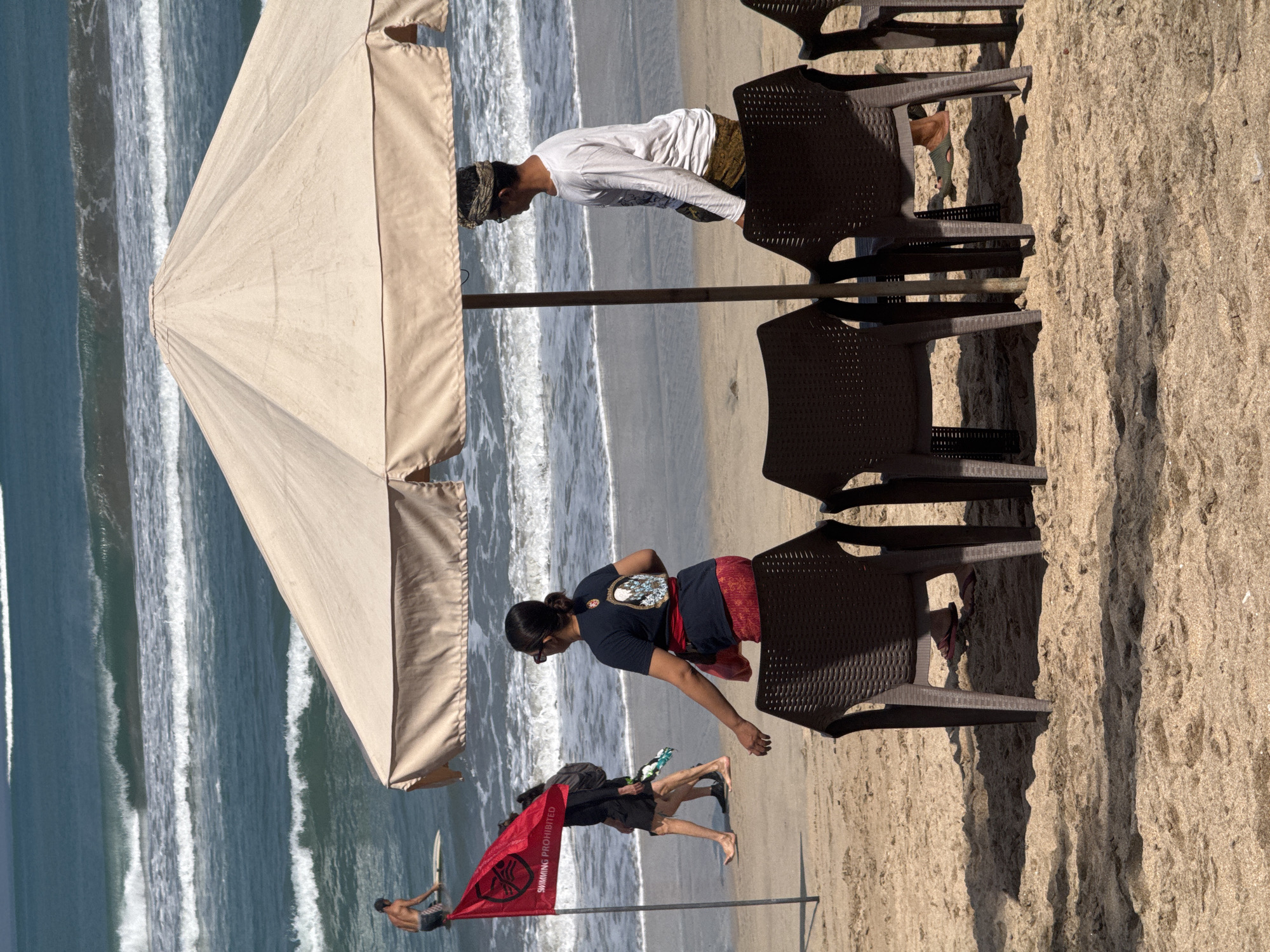 An overcast beach scene in Kuta, Indonesia, is depicted, with the ocean and waves in the background. In the foreground, several brown plastic chairs sit on the sand under a beige beach umbrella. To the left, a red flag with a white symbol and the text "SWIMMING PROHIBITED" is planted in the sand. Three individuals are visible. One, a man with a surfboard, is in the water near the shore. Two other individuals are walking on the beach, one behind the other, with one carrying a backpack. A woman is seated in one of the chairs, facing away from the viewer, and a man stands near the umbrella, looking out at the ocean. The sandy beach is textured with shadows cast by the umbrella and chairs.