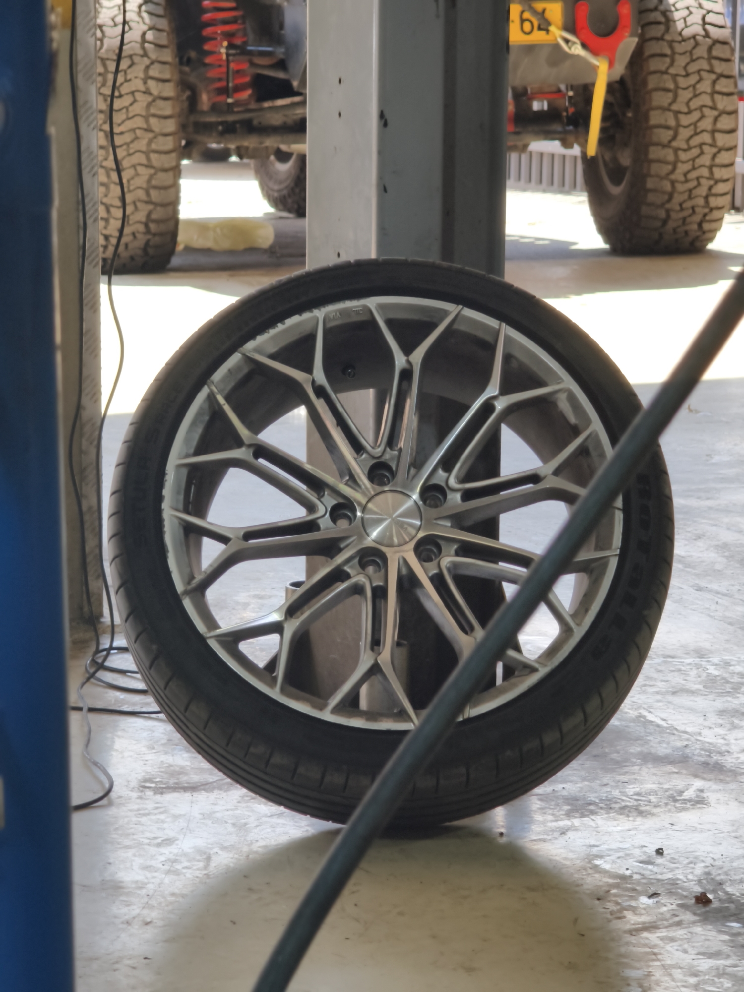In Kafr Yasif, Israel, a close-up, eye-level, outdoor shot captures a car wheel and tire in focus, set against a blurred background of a garage or workshop. The wheel is a complex, multi-spoke design in a metallic silver finish. The tire is black, with visible tread patterns. The ground beneath the wheel appears to be concrete, with some scuff marks and debris. In the background, the rear of a vehicle is partially visible, featuring a large, knobby tire and red suspension components. A yellow license plate with the number "64" is also visible on the rear of the vehicle. Electrical cables snake across the ground. The overall lighting suggests daytime.
