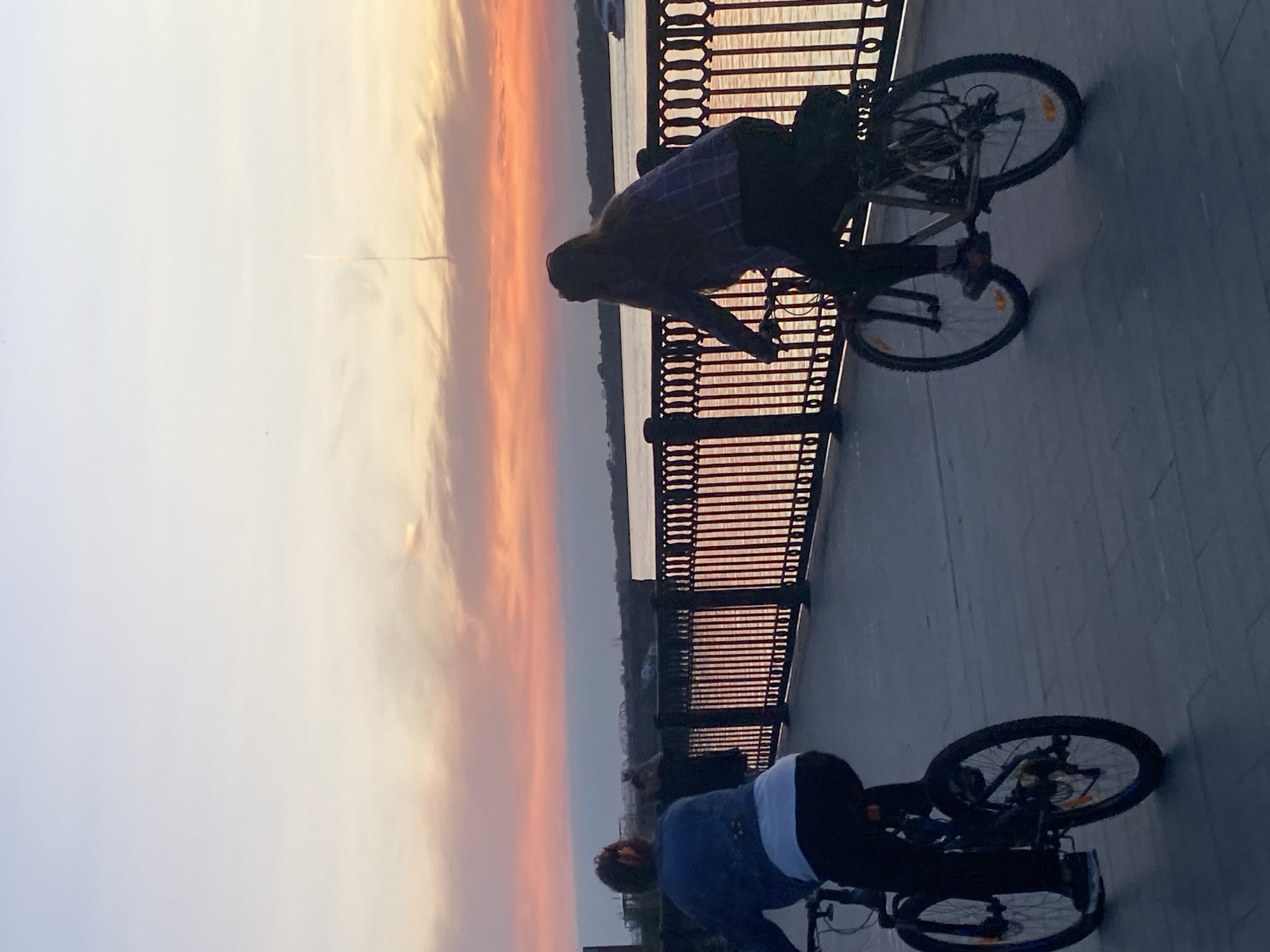Two people are cycling on a paved walkway next to a river at sunset. The sky is filled with vibrant orange and pink hues, indicating a dramatic sunset. A decorative metal fence separates the walkway from the river. The person in the foreground on the right is wearing a plaid shirt and dark pants, while the person on the left is wearing a blue denim jacket and dark pants. Both are riding mountain bikes. The walkway appears to be part of a promenade or embankment, possibly along the Kama River in Perm, Russia, given the characteristic sunset colors and riverfront setting often seen in the city.