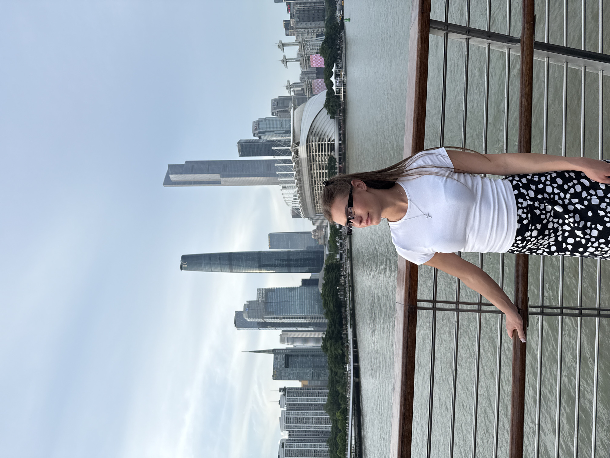 In the Haizhu District of Guangzhou, China, a young woman stands on a wooden promenade overlooking the Pearl River. The woman, with her long brown hair pulled back, wears a white t-shirt and a black and white spotted skirt. She has glasses on and is looking to her left, with her left hand resting on the railing. The cityscape behind her is dominated by modern skyscrapers, including the IFC tower and the Canton Tower, and a large stadium. The sky is overcast, and the water of the river is a muted green.