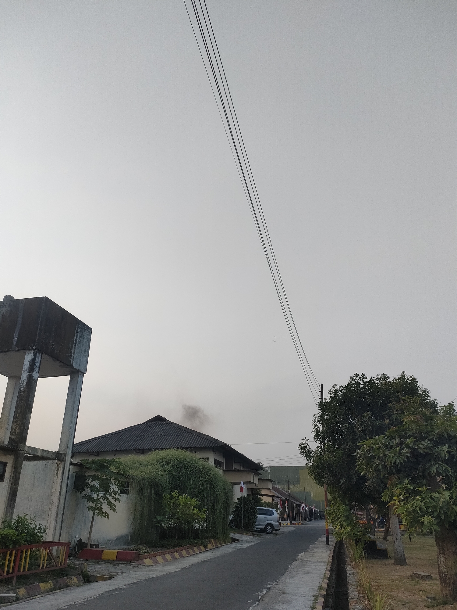 The image shows a street scene in Banguntapan, Indonesia. On the left, a concrete water tower stands next to a building. The building has a dark, sloped roof and is partially covered in lush, trailing green vegetation. A narrow road, lined with a concrete curb and planters, leads into the distance. Several cars are parked along the street, and some buildings with flags are visible on the right side of the road. Tall trees with dense foliage line the right side of the street, casting shadows on the ground. Power lines run diagonally across the pale, overcast sky. A plume of dark smoke is visible rising from behind the roof of the building on the left. The overall impression is of a quiet, residential street in a somewhat hazy or overcast environment.