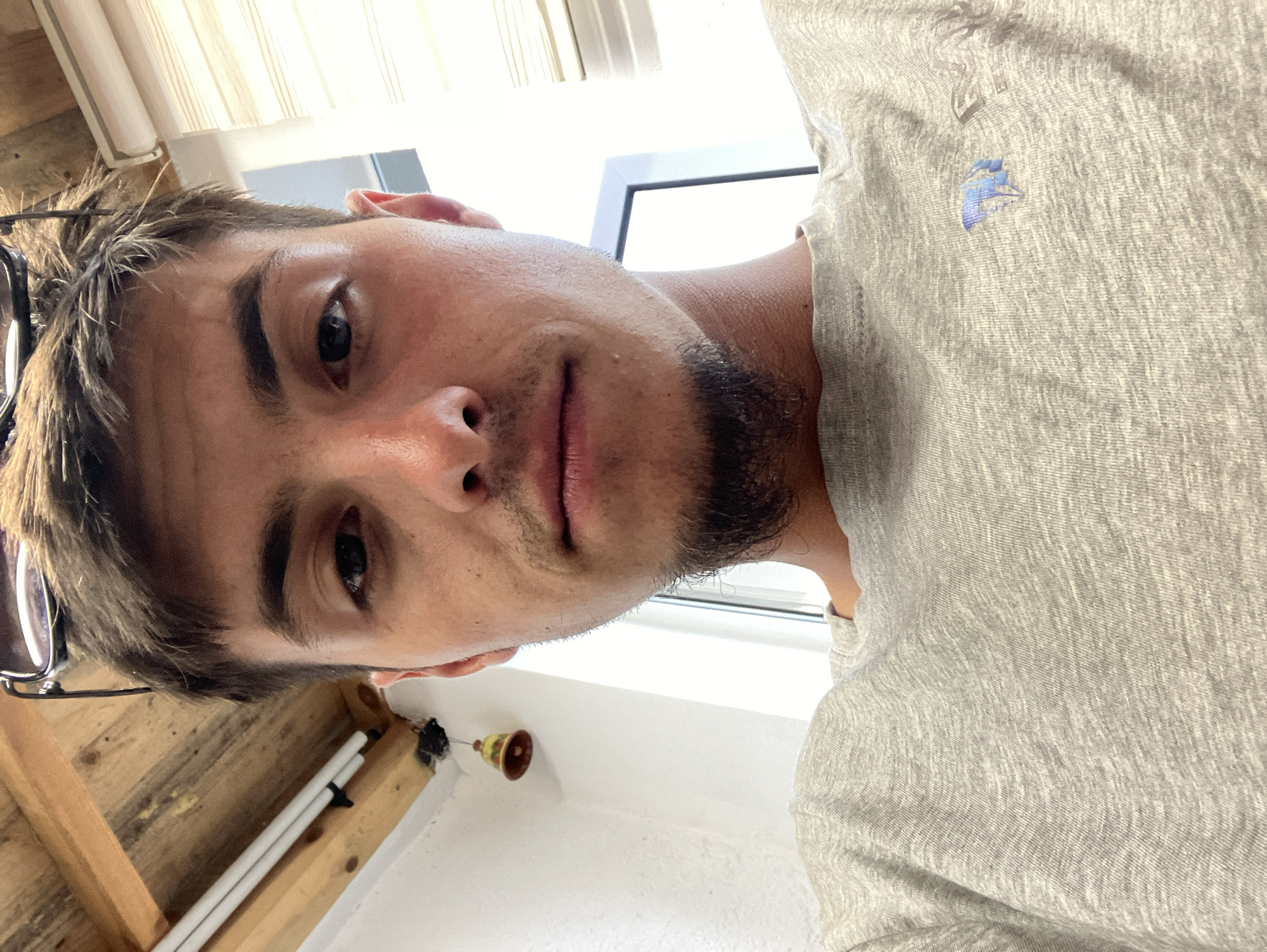 A close-up selfie of a Caucasian male with short brown hair, stubble beard, and visible pores on his face, wearing a light grey t-shirt with a small logo on the chest. Part of a window and a light fixture are visible in the background. The scene is likely indoors, with wooden beams and pipes suggesting construction or a partially finished space. The location in Komuniga, Bulgaria, cannot be determined with the given information.