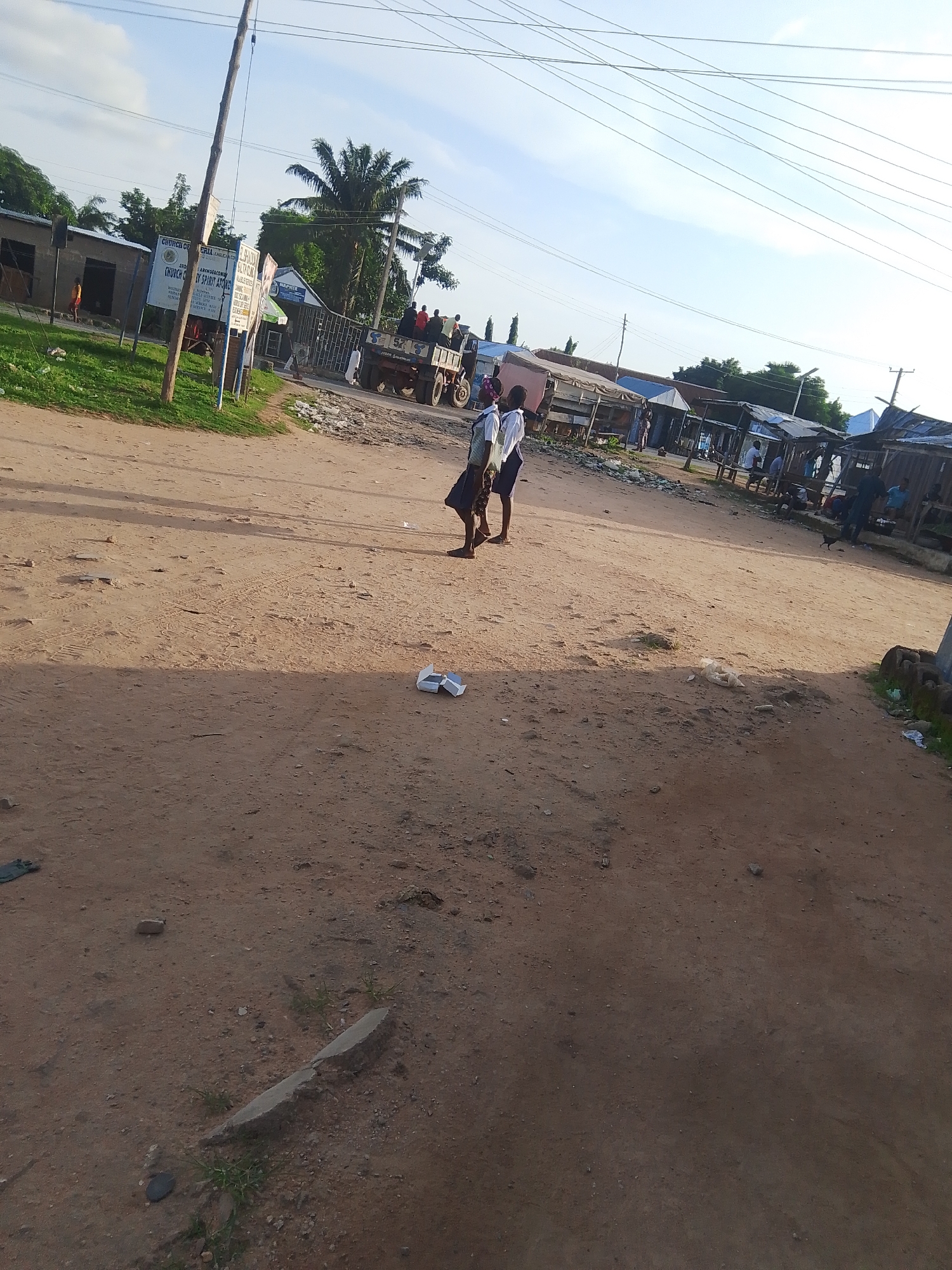 The image depicts a daytime scene in Jalingo, Nigeria. The foreground is dominated by a dusty, unpaved road with tire tracks visible. In the middle ground, two young people in school uniforms, a boy and a girl, are walking on the road. The boy is wearing a white shirt and blue shorts, while the girl is in a white blouse and a blue skirt. Behind them, a truck is parked, and several people are gathered around it.
To the left of the road, there are some buildings, including what appears to be a small structure with a grass-covered roof. A wooden pole with electrical wires stretches across the sky. A signpost is visible on the left, featuring text that includes "CHURCH OF NIGERIA" and "ARCHBISHOP ST PETRO". Further down the road, there are more makeshift structures that seem to be shops or market stalls, with people milling about.
The sky is bright and appears to be a clear, sunny day, suggesting it is likely late morning or early afternoon. The overall setting is a rural or semi-urban area characterized by simple infrastructure and a dusty environment. The presence of school children and people around a truck suggests a normal day with activities like school attendance and local transport or trade.
