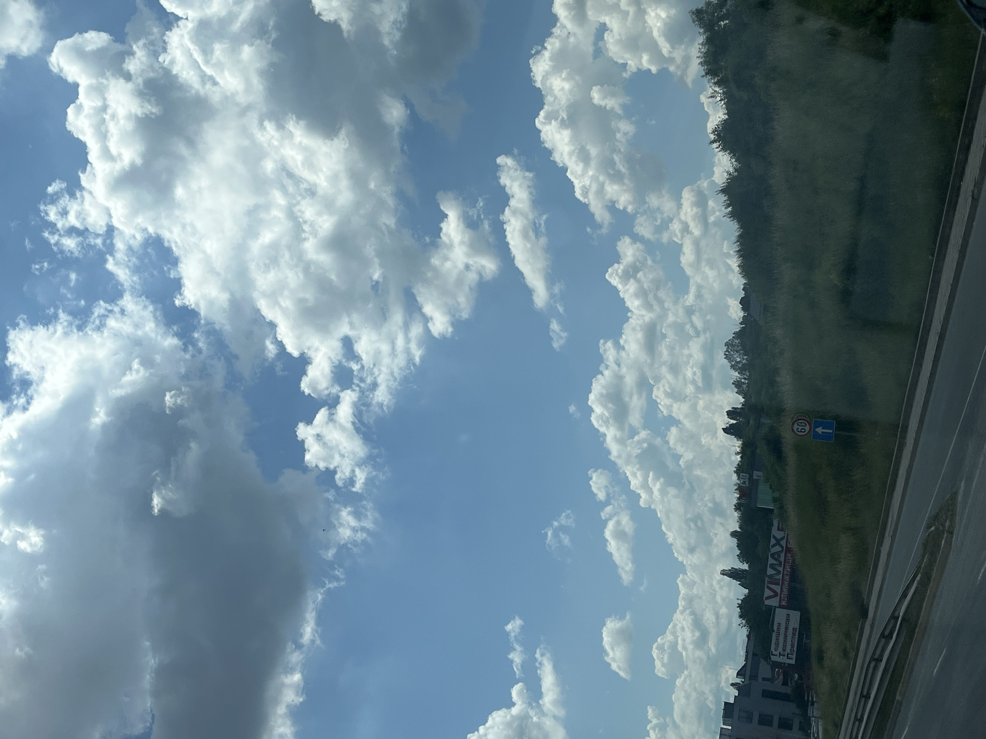 The image shows a wide shot of a suburban or rural landscape under a partly cloudy sky. In the foreground, there is a road with a guardrail. Beyond the road, there is a grassy slope with some trees at the top. On the left side of the slope, there are buildings and a large billboard with text that appears to be in Cyrillic and Latin alphabets. One part of the billboard says "VIMAX" and another part says "Годишен Технически Преглед" which translates to "Annual Technical Inspection". There are also some traffic signs visible. The sky is filled with large, white, fluffy cumulus clouds against a bright blue background. The overall scene suggests a sunny day in a location with some commercial activity.