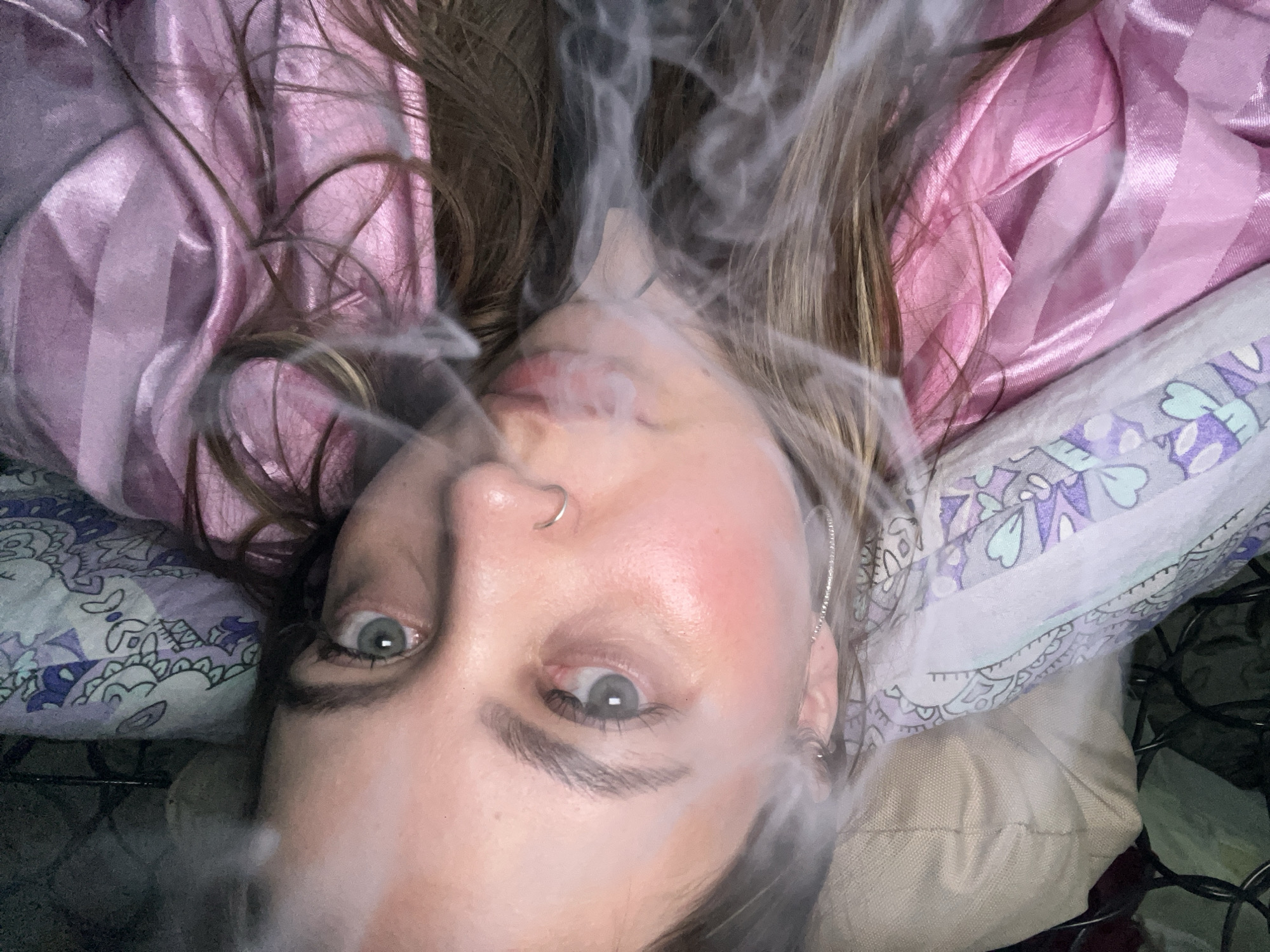 A young woman with light skin and green eyes is lying down, exhaling smoke. She has a nose ring and a silver earring. Her hair is light brown with blonde highlights. She is wearing a pink satin shirt and is surrounded by pillows with floral patterns. The environment is dimly lit. There is no information to suggest the location is in Novopokrovskaya, Russia.