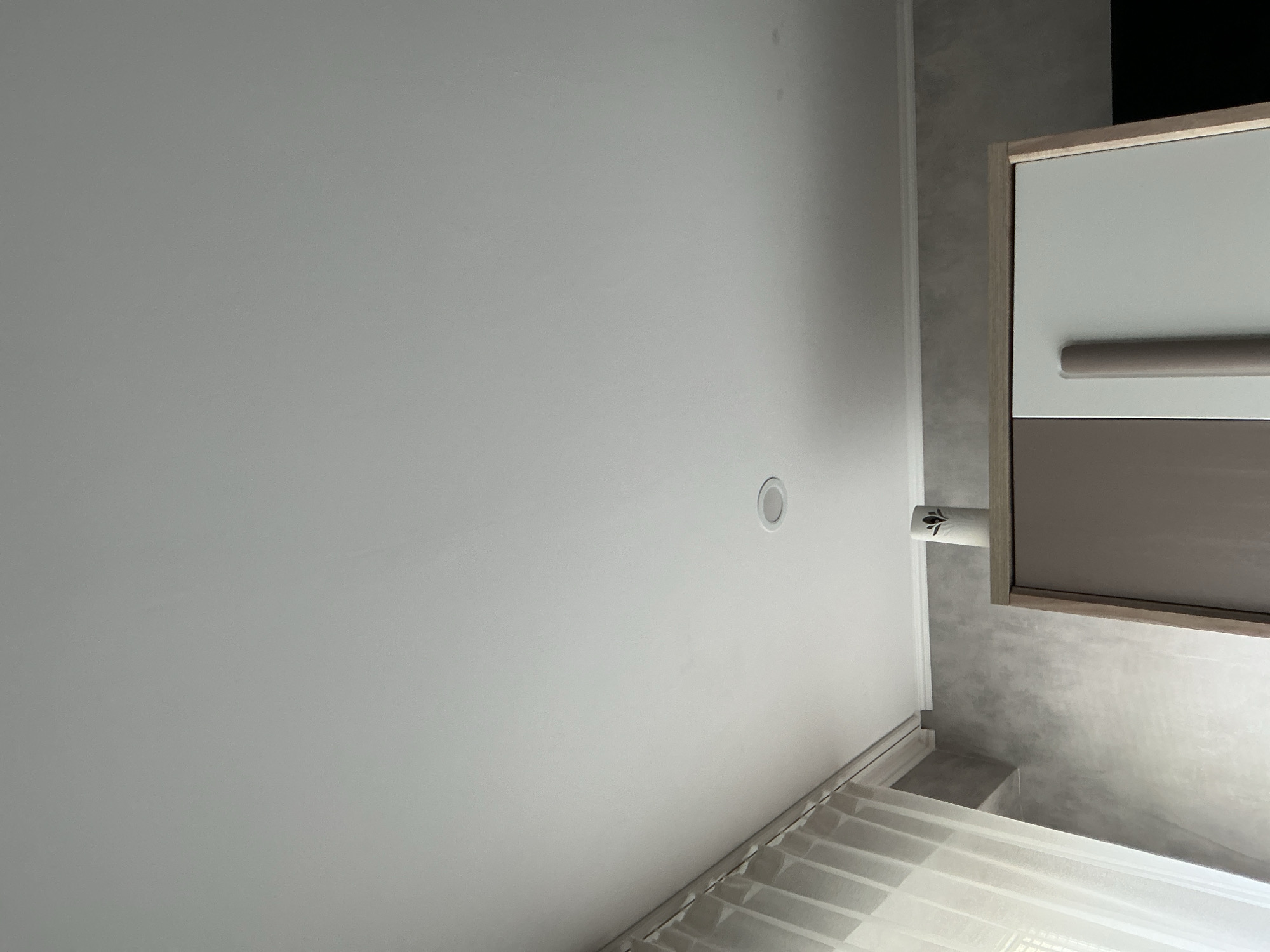 The image shows a room with a focus on a white ceiling with a recessed light fixture. Part of a cabinet is visible on the right, featuring a two-tone design with white and brown sections. There is a handle on the upper section. The background wall is gray. Blinds are seen in the bottom corner. No people are visible. The location is Lovech, Bulgaria.