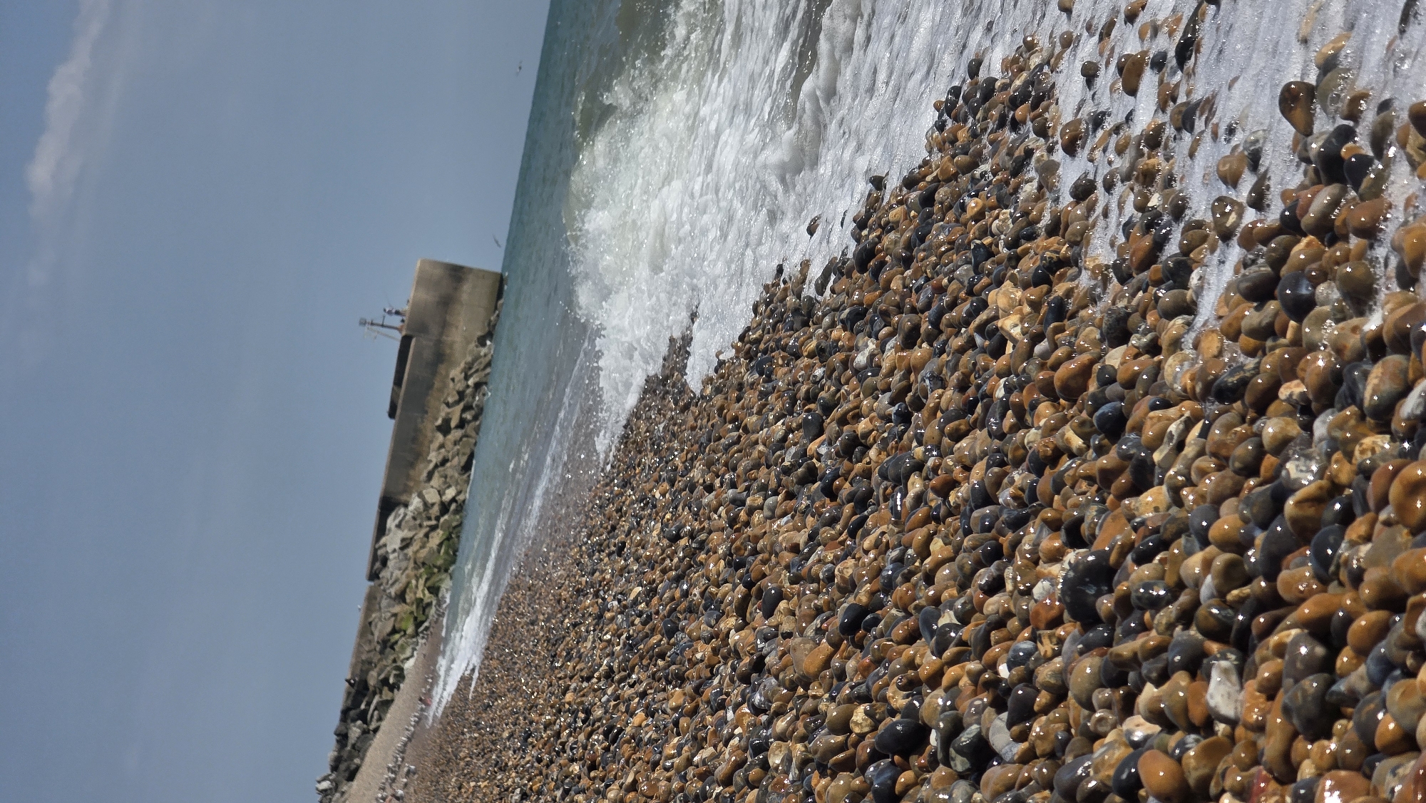 This is a photograph of a shingle beach at St Leonards, United Kingdom. The foreground is dominated by a dense collection of smooth, multi-colored pebbles, some wet and glistening from the incoming tide. Gentle waves with white foam are washing over the lower edge of the shingle. In the mid-ground, a concrete and rock breakwater extends into the sea. A small structure, possibly a navigational marker or a light, is visible on the far end of the breakwater, along with at least two small figures, one appearing to be standing. A few distant birds are visible in the air over the sea. The sky is a pale blue with a wispy cloud formation towards the upper right. The image is taken from a low angle, looking slightly upwards towards the breakwater and sky.