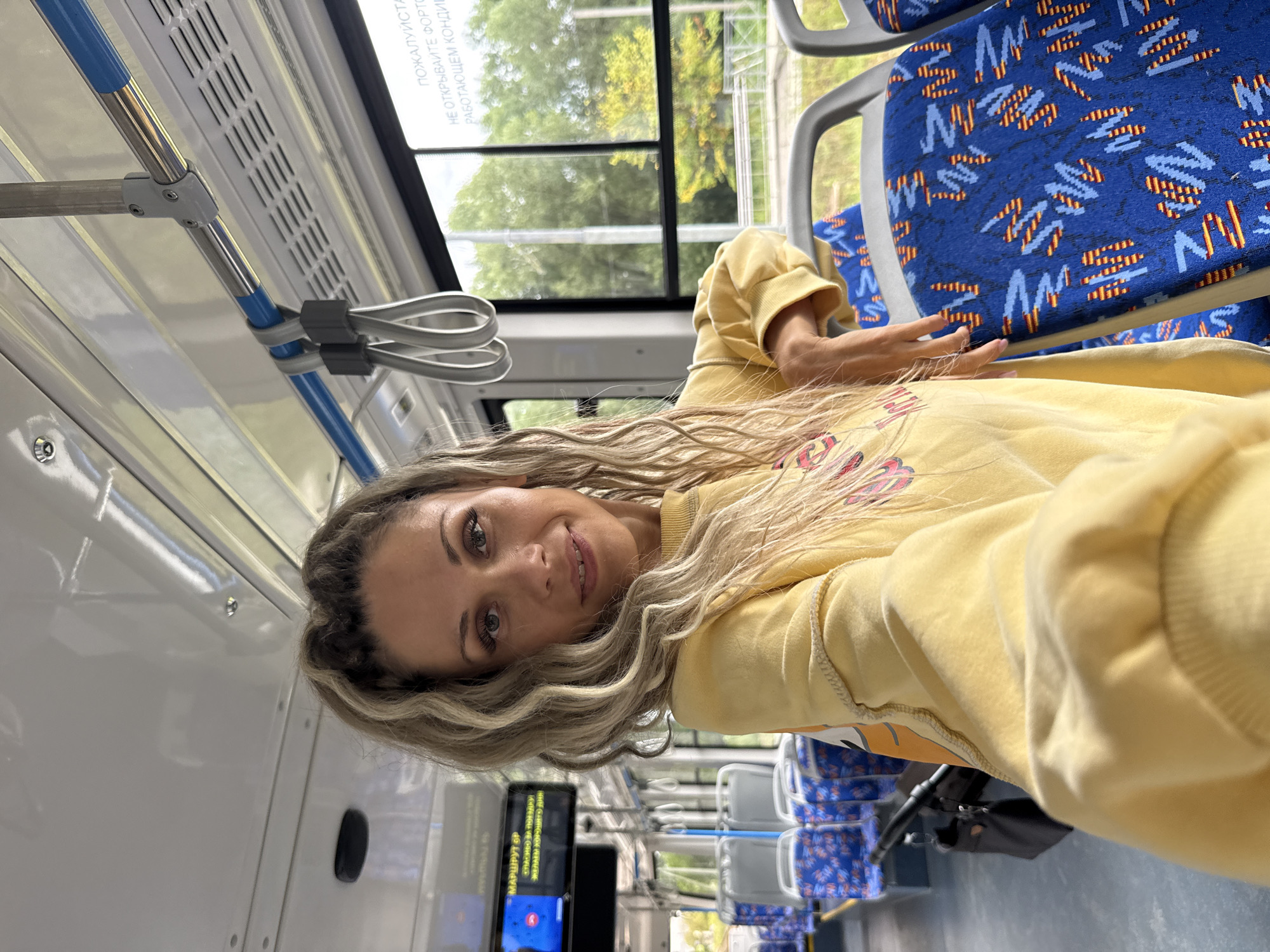 A woman with blonde, wavy hair is taking a selfie inside a public transport vehicle. She is wearing a yellow sweatshirt and has her hand resting on a blue and patterned seat. The interior of the vehicle features rows of seats with blue upholstery, metal poles, and hand straps. Through the windows, trees and greenery are visible, suggesting an outdoor environment. A digital display board with text is mounted on the wall of the vehicle. The scene appears to be set during the daytime, with natural light filtering into the cabin.