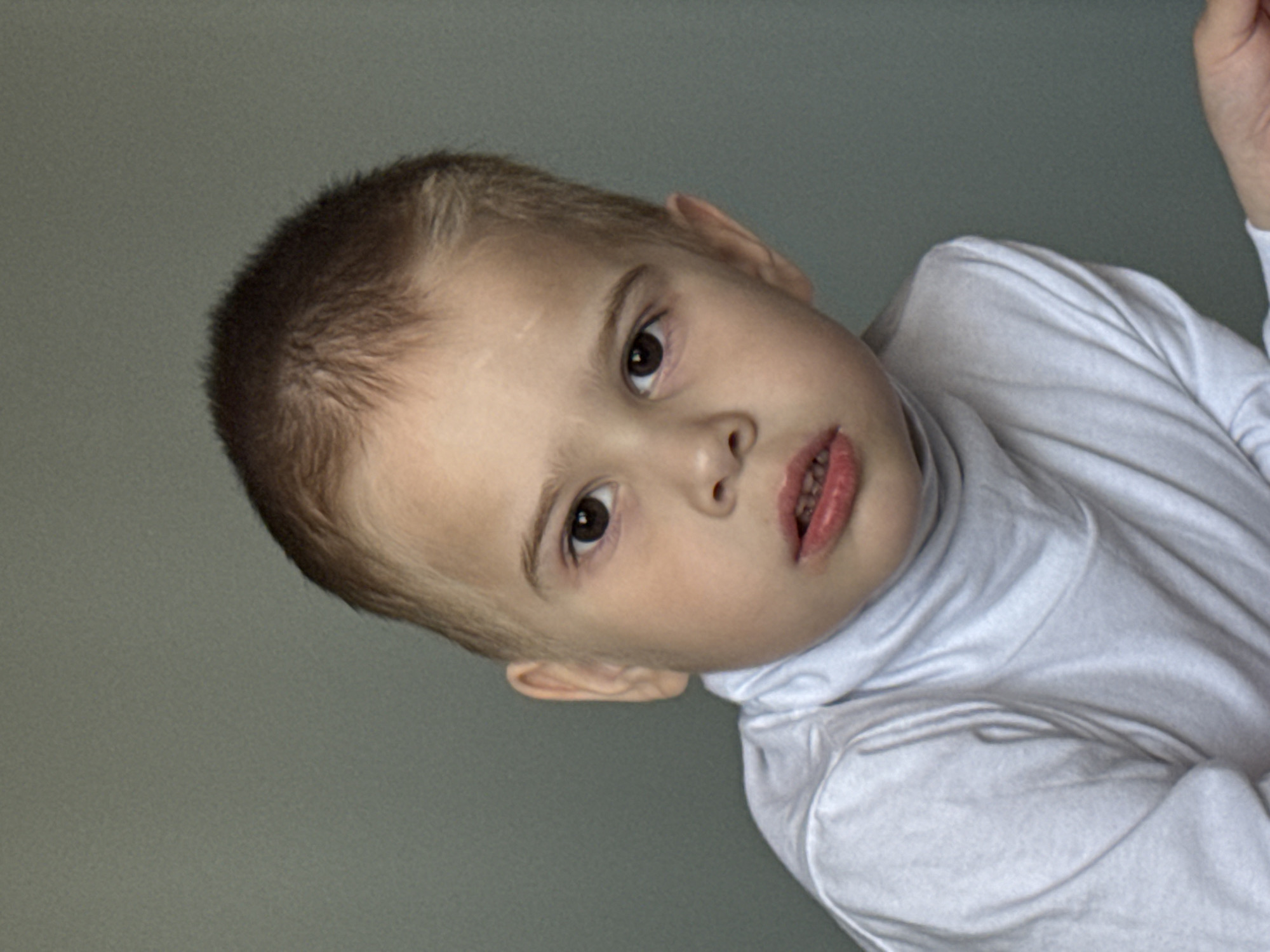 A young child with fair skin and short brown hair is depicted. The child's face is turned slightly to the side, and the child is wearing a white turtleneck shirt. There is a faint scar visible on the child's forehead. The background is a solid, neutral gray. The photograph appears to have been taken indoors. There is no information to identify the location in Chaikovsky, Russia.