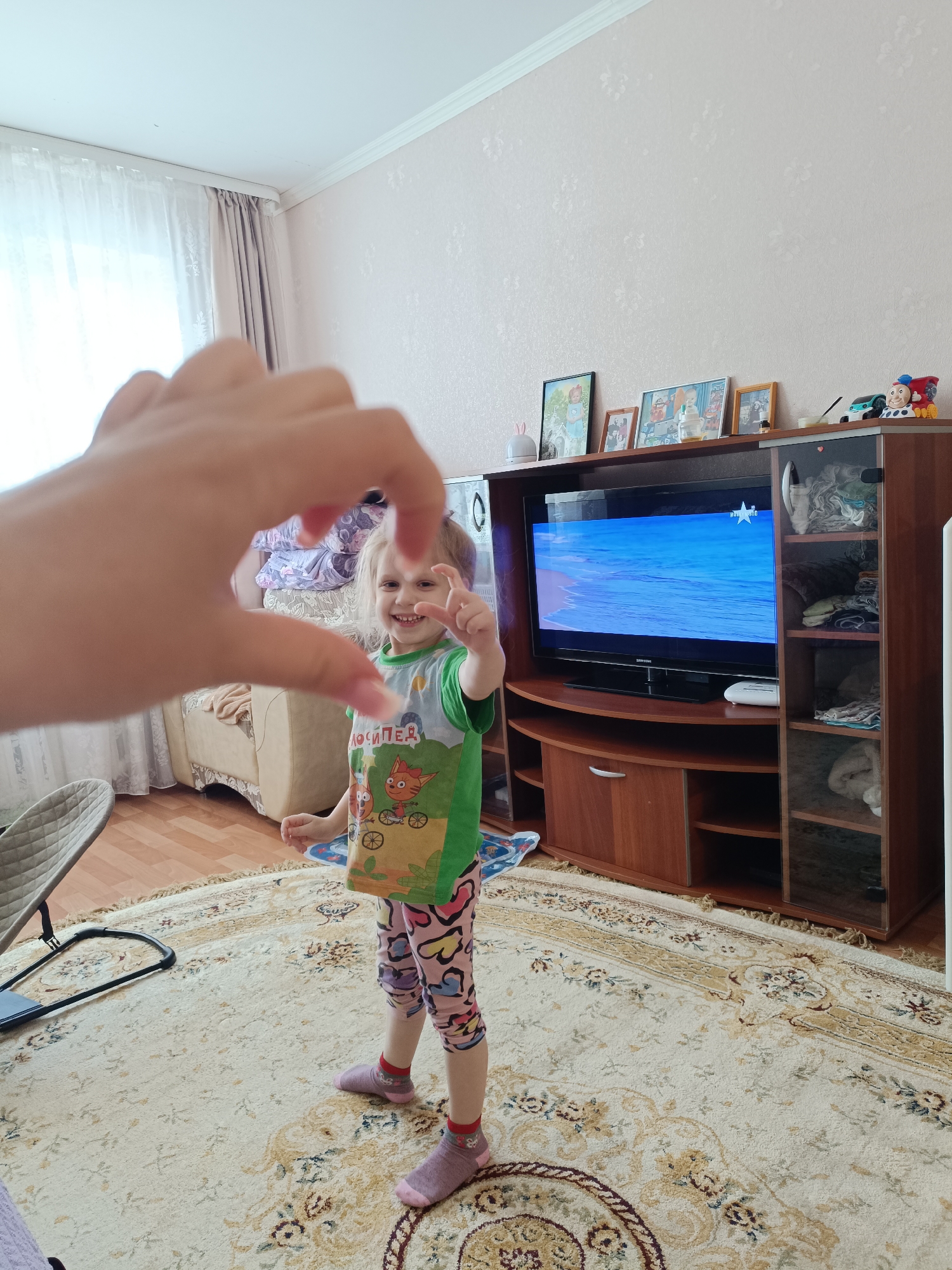 A young child stands on a patterned rug in a room with pink walls. The child, wearing a green shirt and pink heart-patterned leggings, smiles and makes a small heart shape with their fingers. A large hand in the foreground also forms a heart shape. Behind the child is a television playing a scene of a blue ocean, housed in a wooden entertainment center. Various framed pictures and decorative items sit on top of the entertainment center. The room is furnished with a beige sofa visible in the background, and light-colored curtains are drawn in front of a window. Based on the location of similar images and the camera metadata, this image was taken in Tobolsk, Russia.