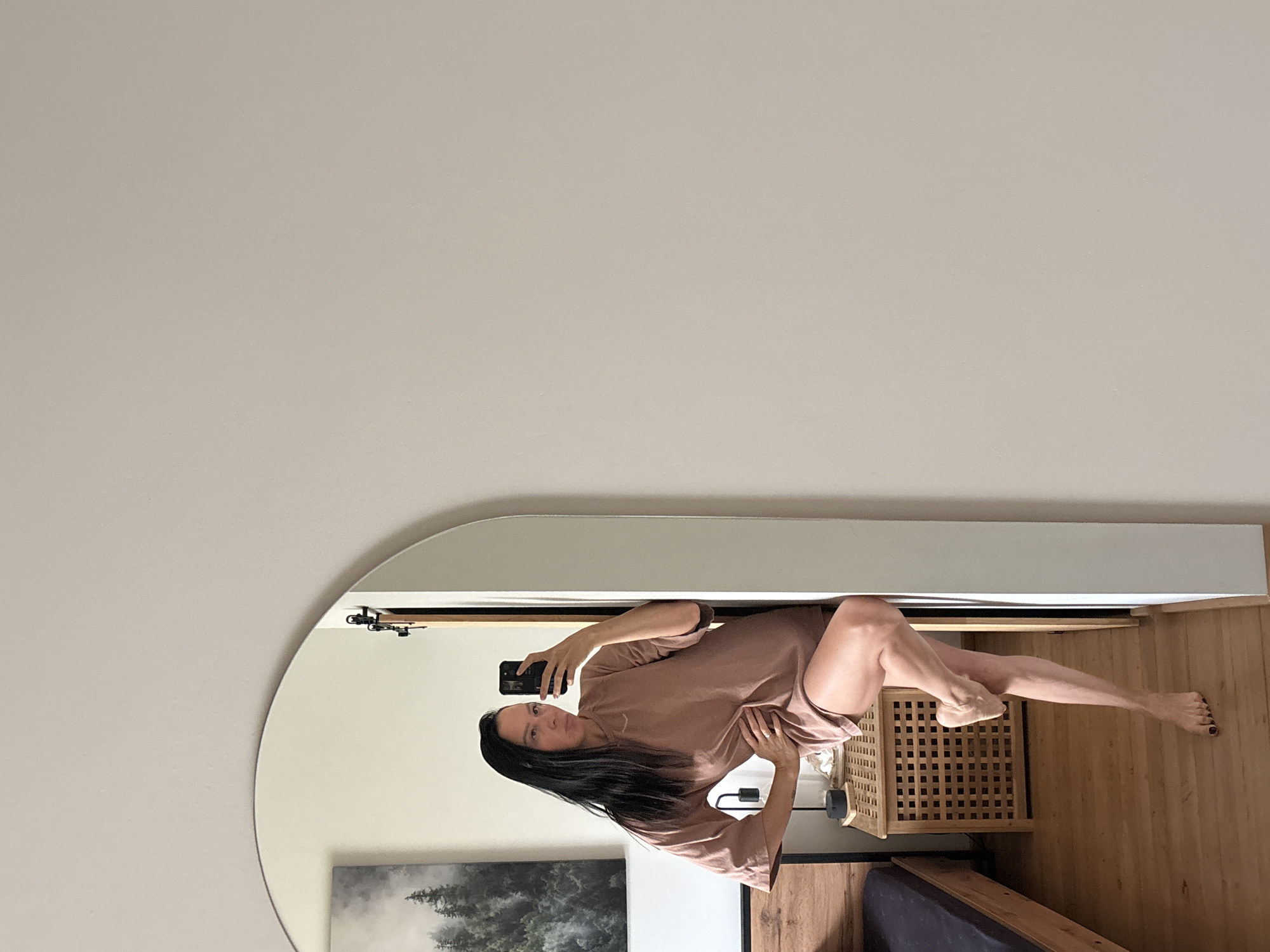 An indoor, full-body mirror selfie shows a woman with long, dark hair wearing a light brown t-shirt dress. She is standing with her right hand on her hip and her left leg bent and raised behind her. She is holding a black smartphone in her left hand, taking the picture. Behind her, a framed artwork of trees in a misty forest hangs on a white wall. To the left of the artwork is a wooden bed frame with a dark mattress. In front of the artwork is a small, cube-shaped wooden nightstand with a grid pattern, on which a black lamp sits. The flooring appears to be wood. The room has a neutral color palette with white walls and warm wood tones. There is no discernible connection to Chaikovsky, Russia, in the image.
