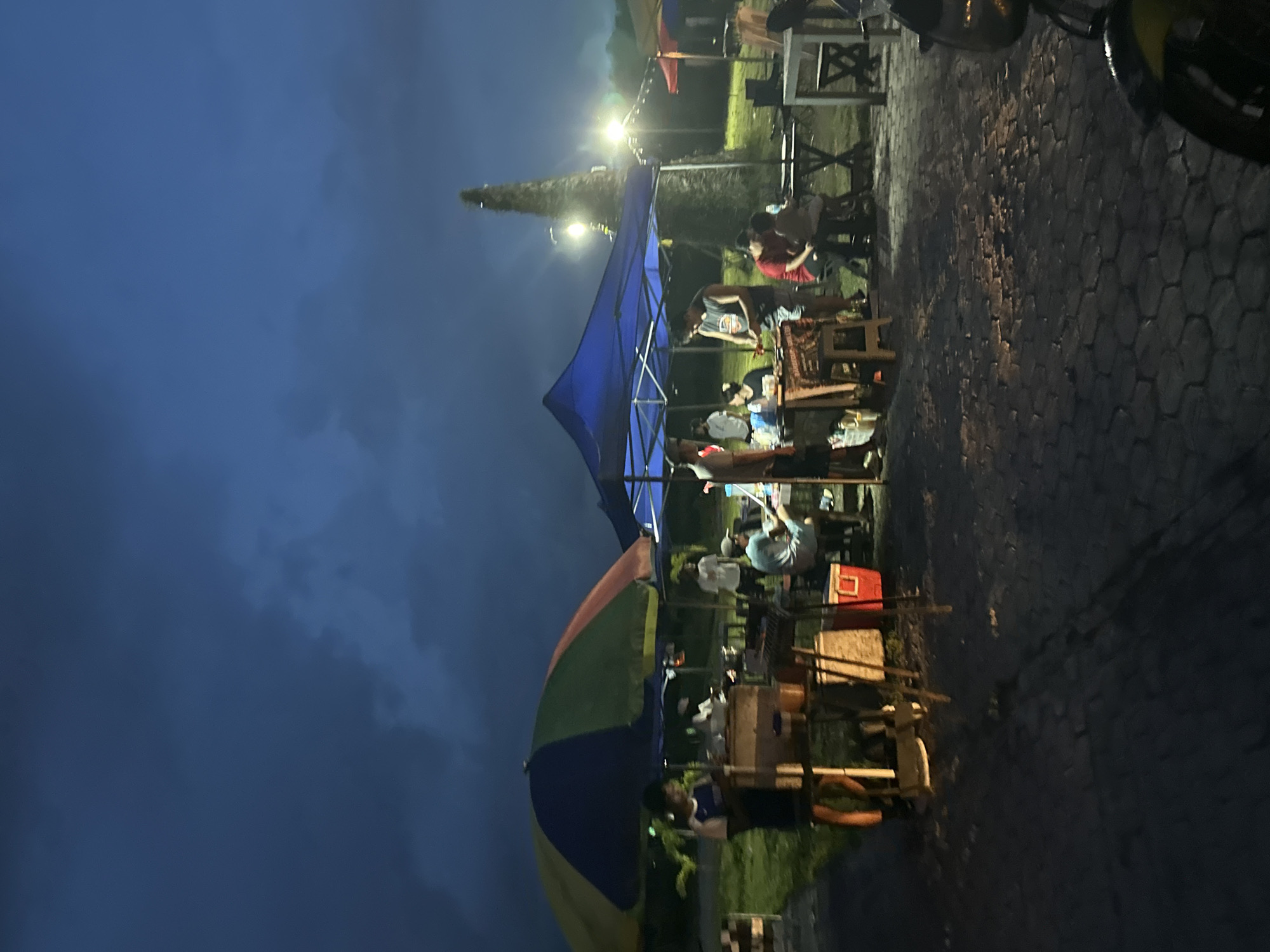 The image depicts a lively outdoor scene at night, likely an informal dining area or market. The dark blue sky, indicative of evening, forms the backdrop for the artificially lit gathering below. The ground is paved with hexagonal stones, and lush green vegetation and tall trees are visible in the background, including a prominent conical tree standing against the dark horizon.
Numerous people are present, mostly seated at simple wooden tables and chairs arranged under canopies and umbrellas. They appear to be socializing, eating, and relaxing in a casual atmosphere. Individuals are dressed in everyday attire, with one person in a red shirt noticeable under a blue canopy.
The setup includes a large blue canopy providing shelter over a cluster of tables, and a vibrant, multi-colored umbrella (featuring blue, green, yellow, and red segments) stands open in the foreground. Several bright artificial lights illuminate the area, casting a warm glow over the scene. A red cooler is visible near some tables. Faint text on a banner or cloth hanging near one table appears to advertise "TUNA BURGER" and "DURIAN KAPE," which are typical food items found in Davao City, Philippines. A small portion of a vehicle, possibly a motorcycle, can be seen in the upper right corner.
The time of day is clearly night, and the weather seems clear without any precipitation. The overall mood is relaxed and social, characteristic of an evening community gathering or food stall experience.