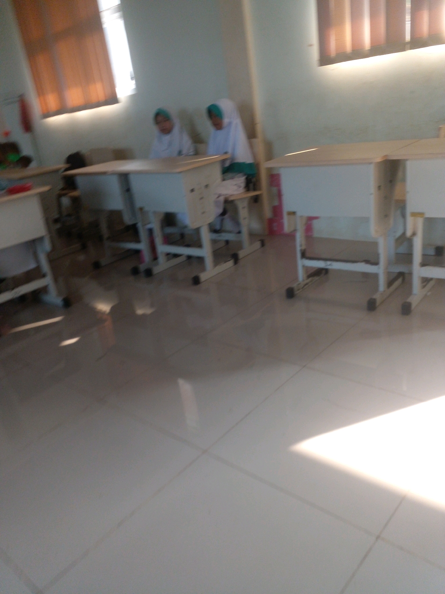 The image depicts an indoor scene, likely a classroom, with tiled flooring and light streaming in from a window covered by blinds. In the background, two young individuals wearing white hijabs and robes are seated at desks. The desks appear to be wooden with white metal legs. The room is sparsely furnished, with additional desks visible in the foreground. The lighting suggests it is daytime. The provided location is Banyuputih, Indonesia.