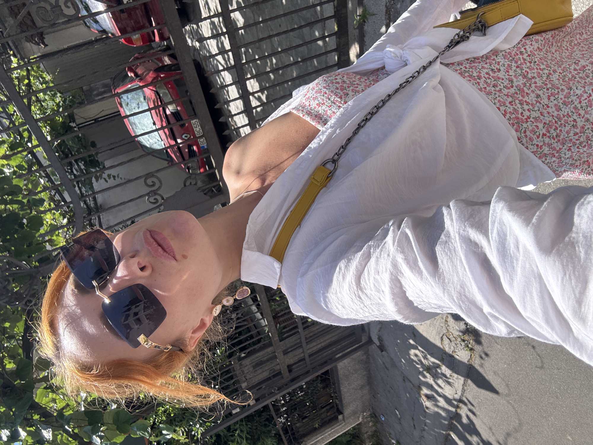 A woman with reddish-blonde hair, wearing large dark sunglasses, a white unbuttoned shirt over a floral print dress, and a yellow crossbody bag, is taking a selfie outdoors. She is standing in front of a black metal fence, behind which two red cars are parked. One car has a license plate that reads "B0 BC 55". The background shows greenery and buildings. The scene appears to be a street in Budva, Montenegro, characterized by its residential architecture and typical Mediterranean vegetation. The lighting suggests it is daytime with bright sunlight.