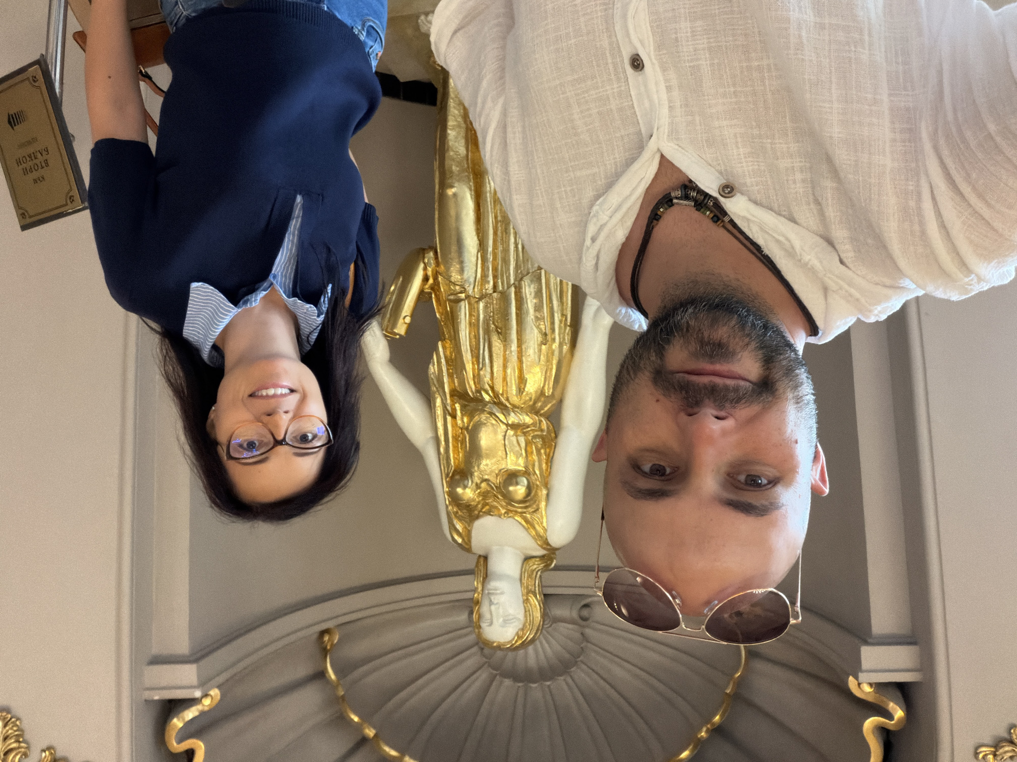 Two individuals, a man and a woman, are posing for a selfie in front of a golden statue of a woman in Sofia, Bulgaria. The man is on the left, wearing a white hooded shirt and sunglasses pushed up on his bald head. He has a beard and is wearing a beaded necklace. The woman is on the right, wearing a dark blue polo shirt with a striped collar and glasses. They are both smiling at the camera. The background shows a decorative wall and the golden statue. A sign with text in Cyrillic script is visible on the right. The setting appears to be a museum or a historical building.