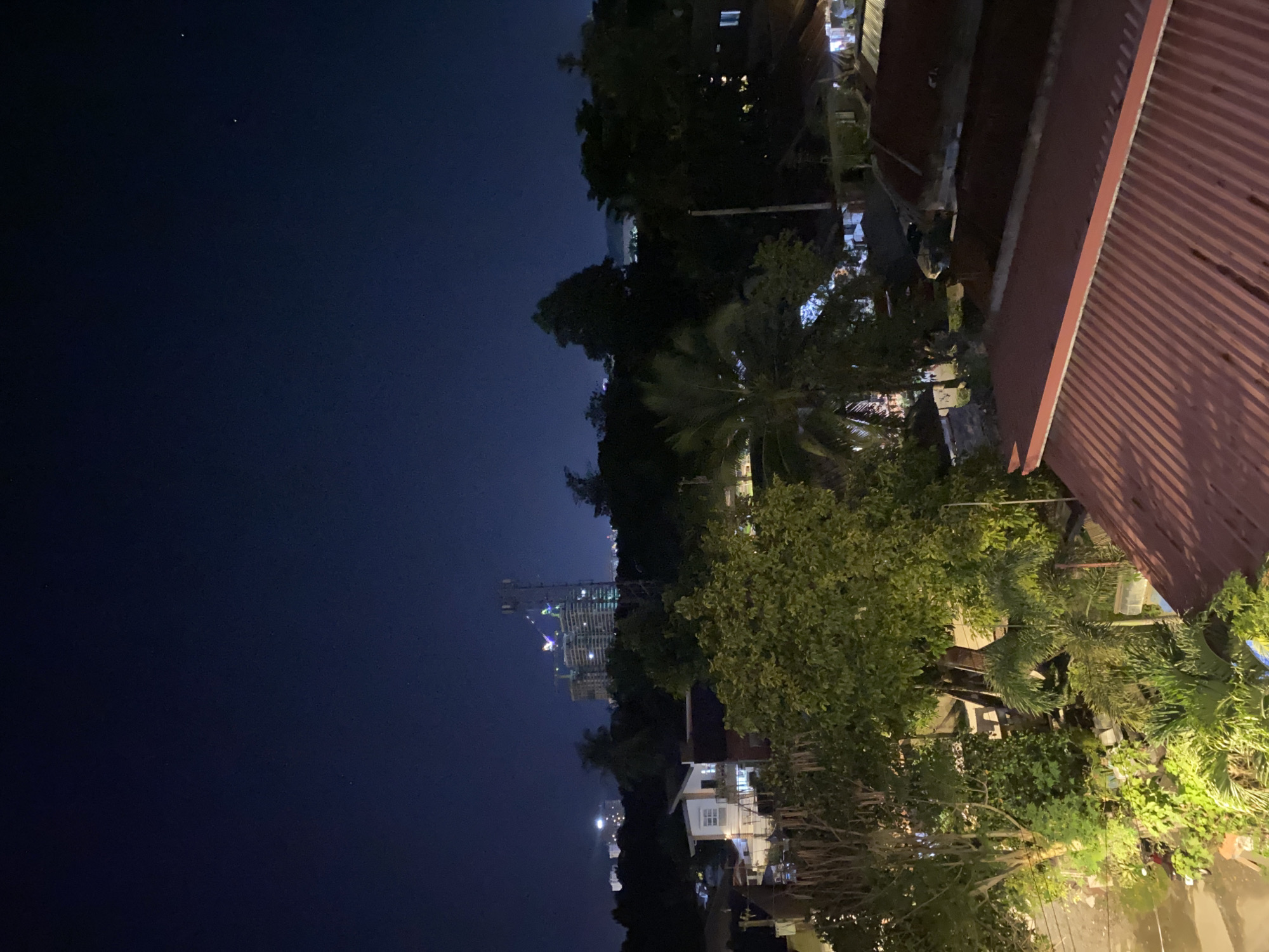 This image captures a wide-angle, elevated night view of a residential and urban landscape in Davao City, Philippines. The dark blue sky, largely devoid of visible clouds, occupies the upper third of the frame, revealing faint stars. In the distant background, a cityscape is visible, characterized by numerous artificial lights. A prominent, multi-story building under construction, illuminated with green and purple lights, stands out, flanked by multiple construction cranes. Adjacent to this structure, a tall cellular tower is clearly discernible.
The midground features dense tropical vegetation, including numerous trees and palm fronds, which partially obscure the view of the terrain below. Scattered among the greenery are several residential structures. One distinct white house with illuminated windows is centrally located, appearing to have two stories.
The foreground is dominated by the reddish-brown, corrugated metal roofs of multiple houses, angled towards the viewer. These roofs exhibit distinct parallel ridges and valleys. Below and to the left of these roofs, a street or pathway is visible, illuminated by artificial light, and a parked motorcycle is faintly discernable. Extensive green foliage and smaller plants are present throughout the midground and foreground, illuminated by various artificial light sources, creating areas of shadow and highlight. No individuals are visible in the scene.