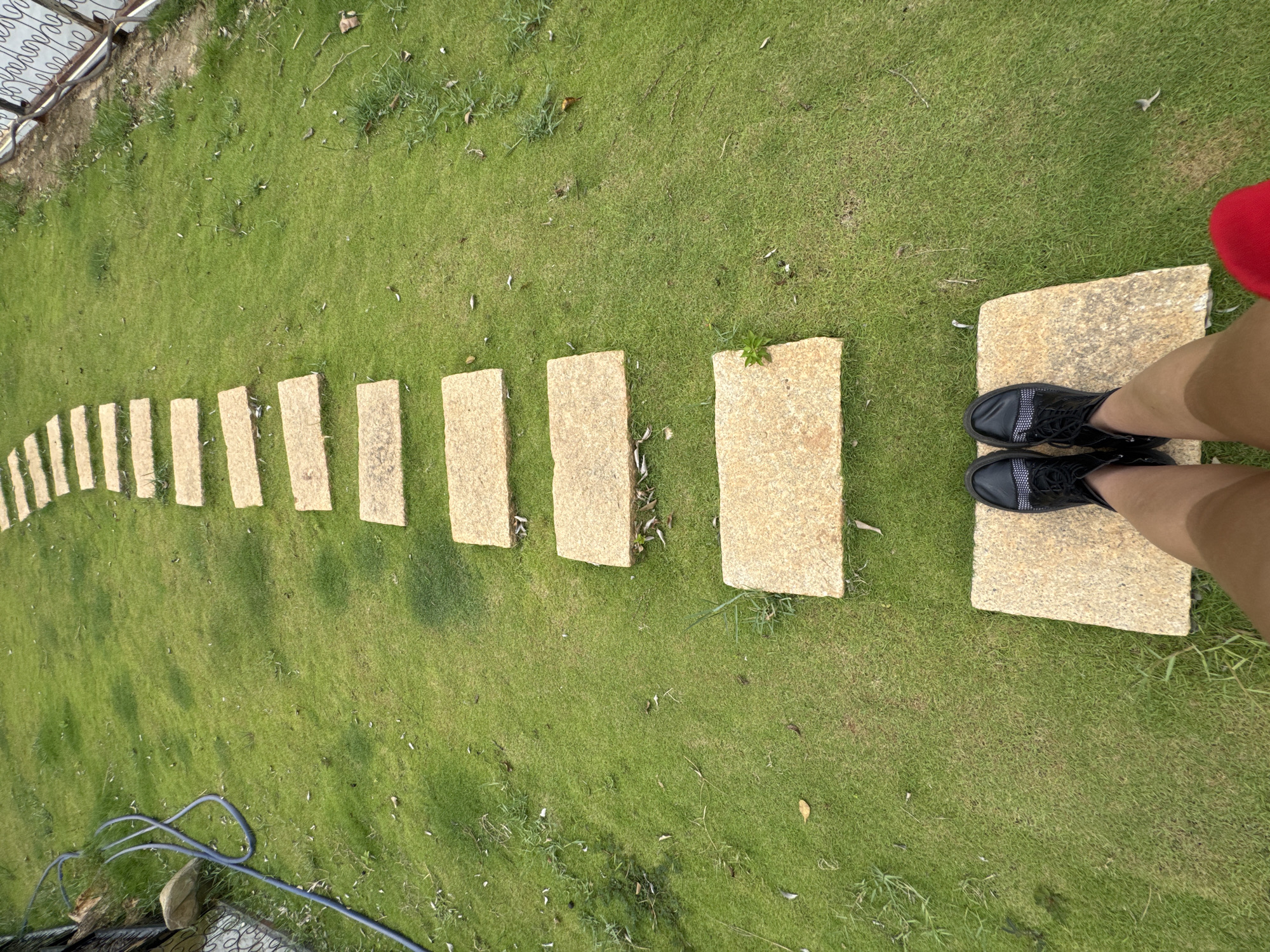 The image shows a path of rectangular stone pavers running through a grassy lawn. The path curves slightly, leading away from the viewer's perspective. At the end of the path, the viewer sees the feet of a person wearing black shoes standing on a pair of the pavers. A piece of red clothing is also visible near the person. The lawn appears well-maintained. A garden hose sits on the grass along the edge of the frame. The location is identified as Phú Thạnh, Vietnam.