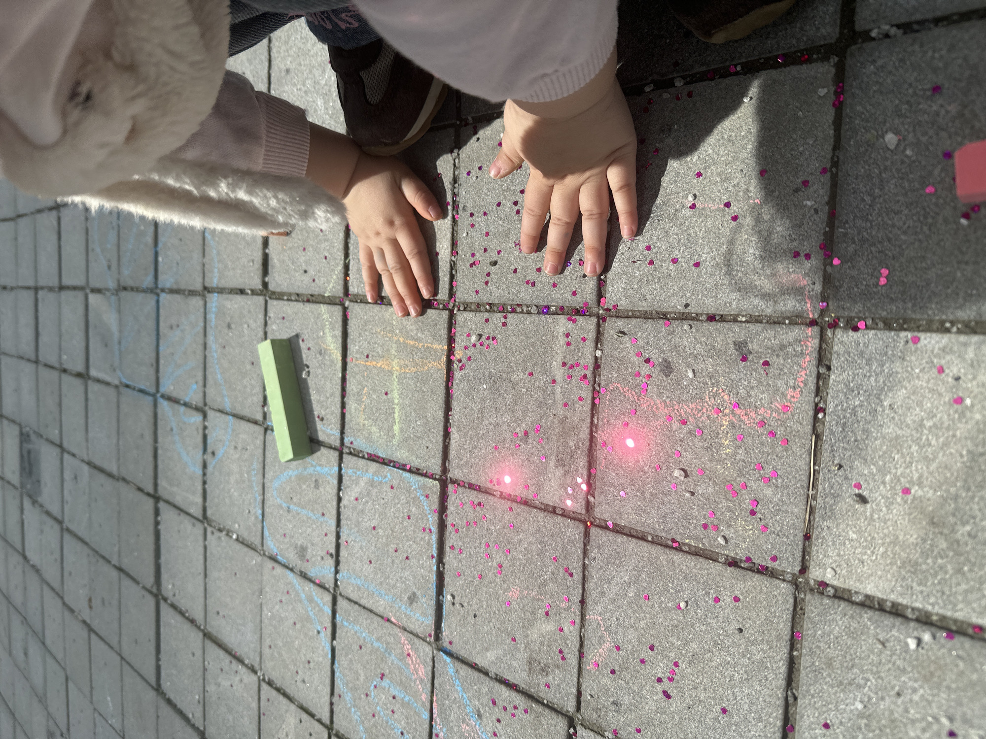 This image, captured at ground level in Yekaterinburg, Russia, features a child interacting with a paved outdoor surface. The ground is composed of grey, square-shaped pavers with visible grout lines. Scattered extensively across these pavers are numerous small, bright pink, heart-shaped glitter pieces, which intensely reflect direct sunlight, creating vibrant specular highlights, particularly in the lower-middle and right sections of the frame. Faint blue chalk lines and scribbles are visible on several pavers, and a solid green piece of chalk rests horizontally on a paver in the upper-right quadrant.
Two small, light-skinned hands, belonging to a child, are placed flat, palms down, on the glitter-strewn pavers in the lower-middle of the image. The child wears a light-colored, long-sleeved garment with ribbed cuffs. A fluffy, white, textured fabric, possibly part of a hood or animal costume, is visible in the upper right. A portion of blue denim jeans and a dark brown shoe with a lighter sole are also visible on the far right. Strong sunlight casts distinct shadows of the child's hands and implied body onto the pavers, indicating bright, clear daytime conditions.